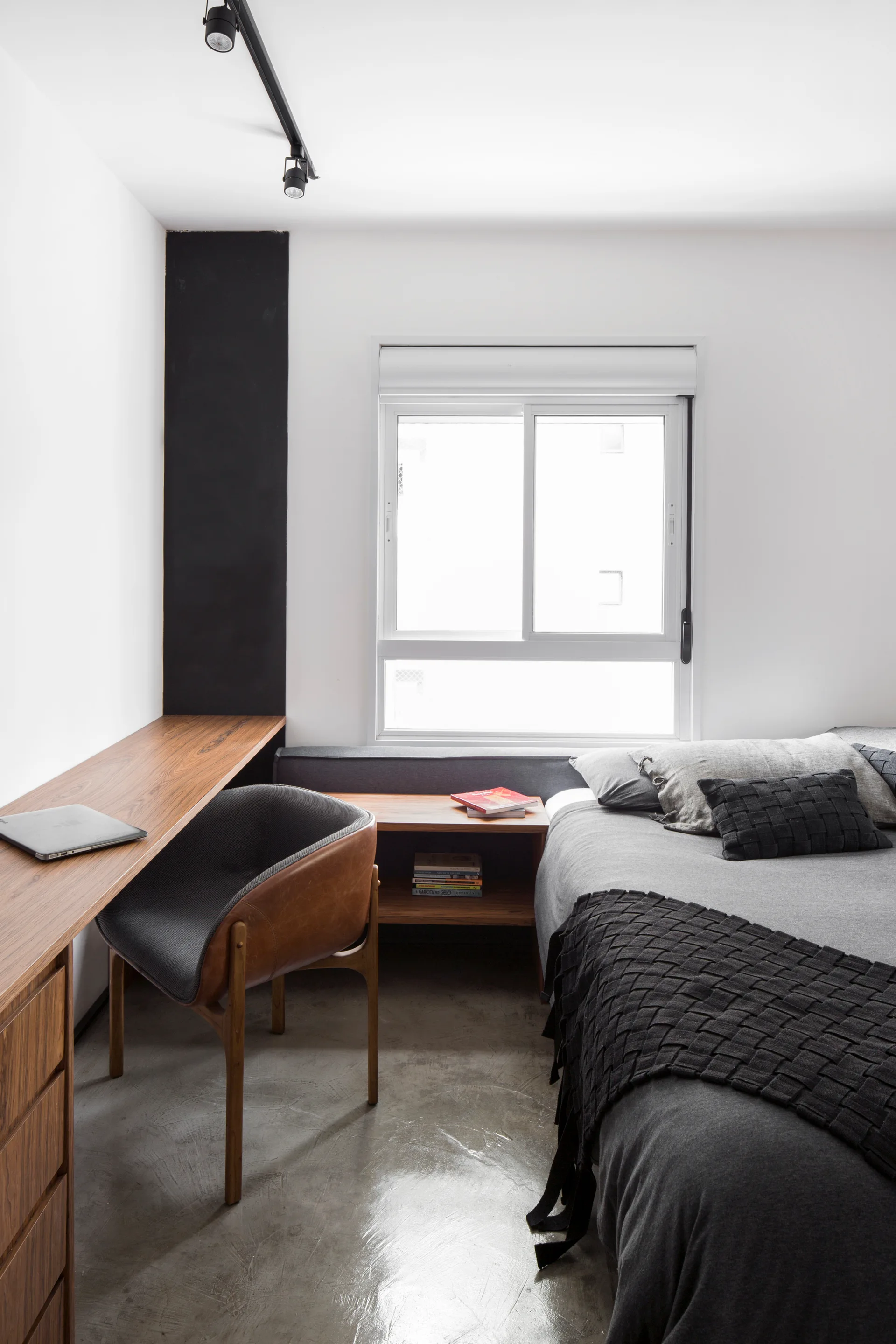 A modern bedroom features a built-in wooden desk with a leather chair, a bed with gray bedding, a polished concrete floor, and track lighting.