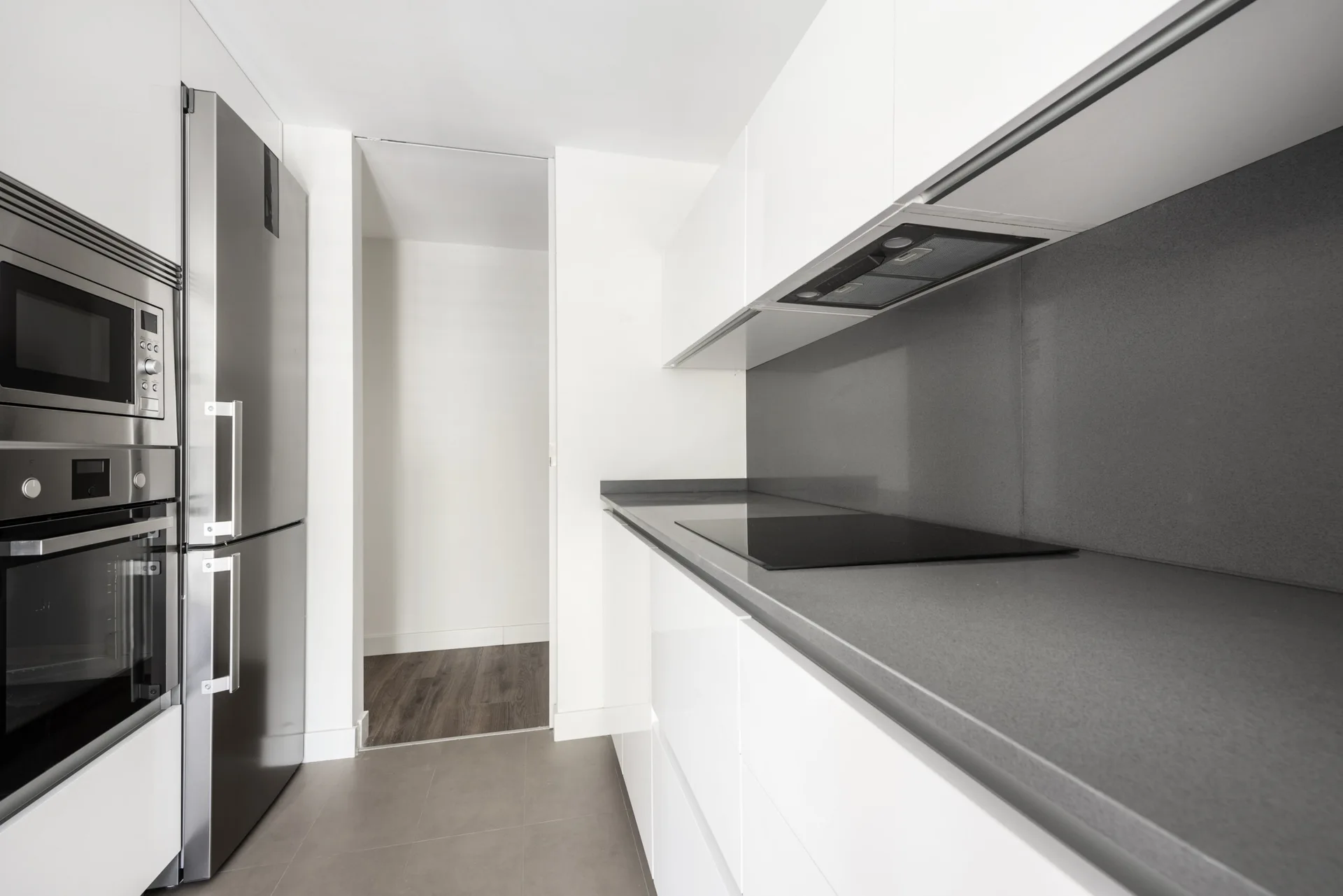 A modern kitchen with handleless white gloss cabinets, a grey stone countertop with matching backsplash, and built-in stainless steel appliances.