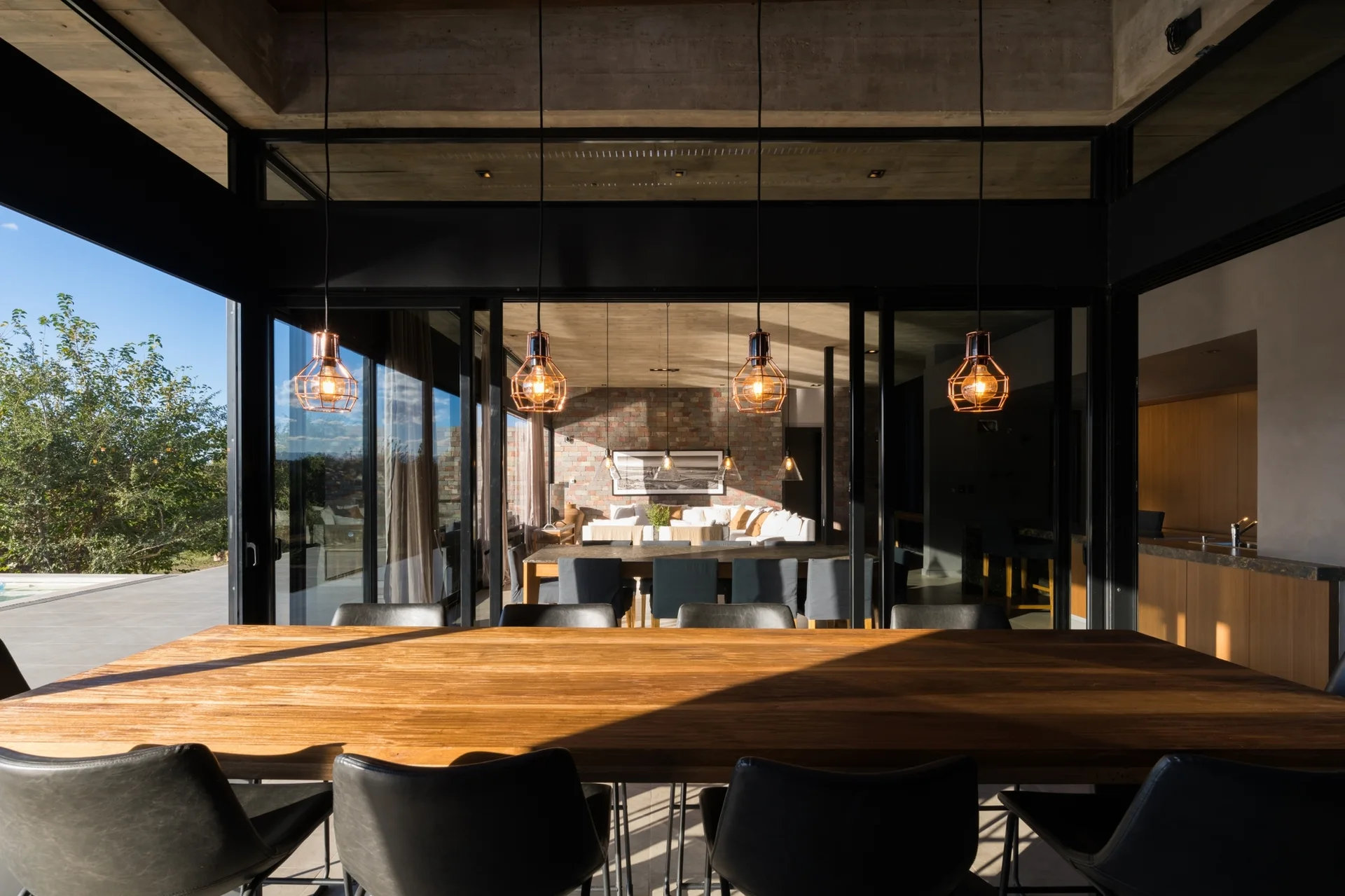 A long wooden dining table with dark chairs sits under four copper cage pendant lights in a room with a concrete ceiling and glass sliding doors.