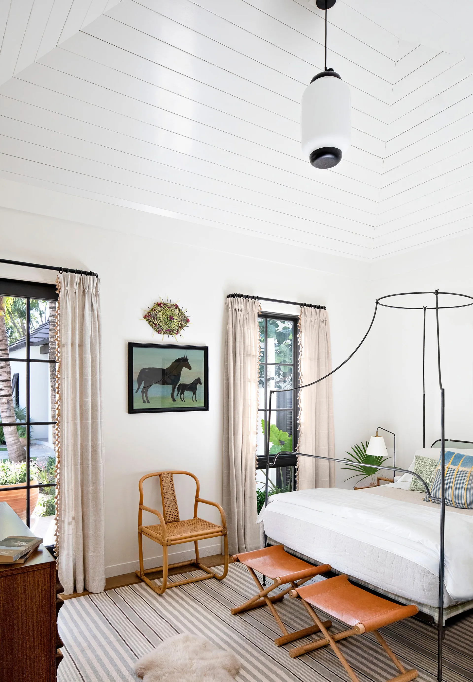 A bright bedroom featuring a black metal canopy bed, leather folding stools, a striped rug, and a white planked vaulted ceiling.