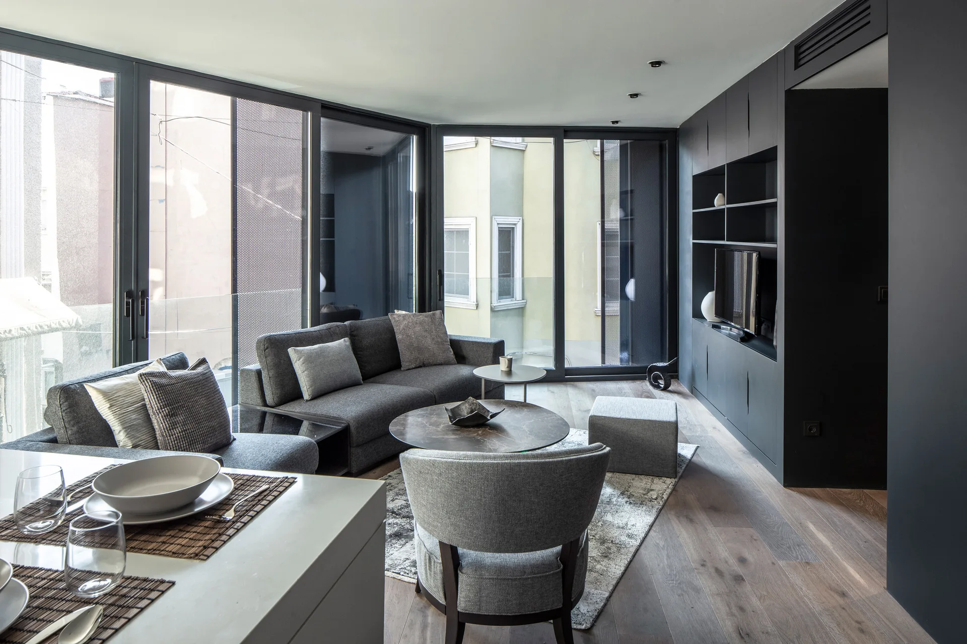 A modern living room features a grey sofa, a curved armchair, and floor-to-ceiling sliding glass doors next to a matte black built-in wall unit.