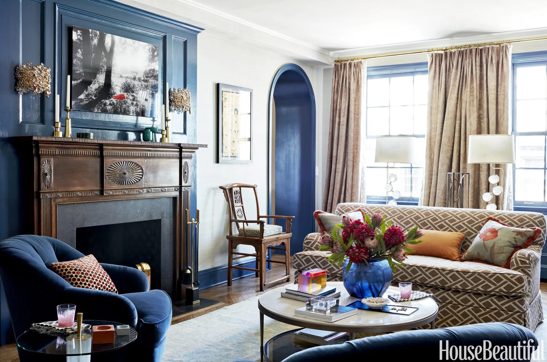 A traditional living room features a dark wood fireplace with navy blue paneled walls, a patterned sofa, a velvet armchair, and an arched doorway.