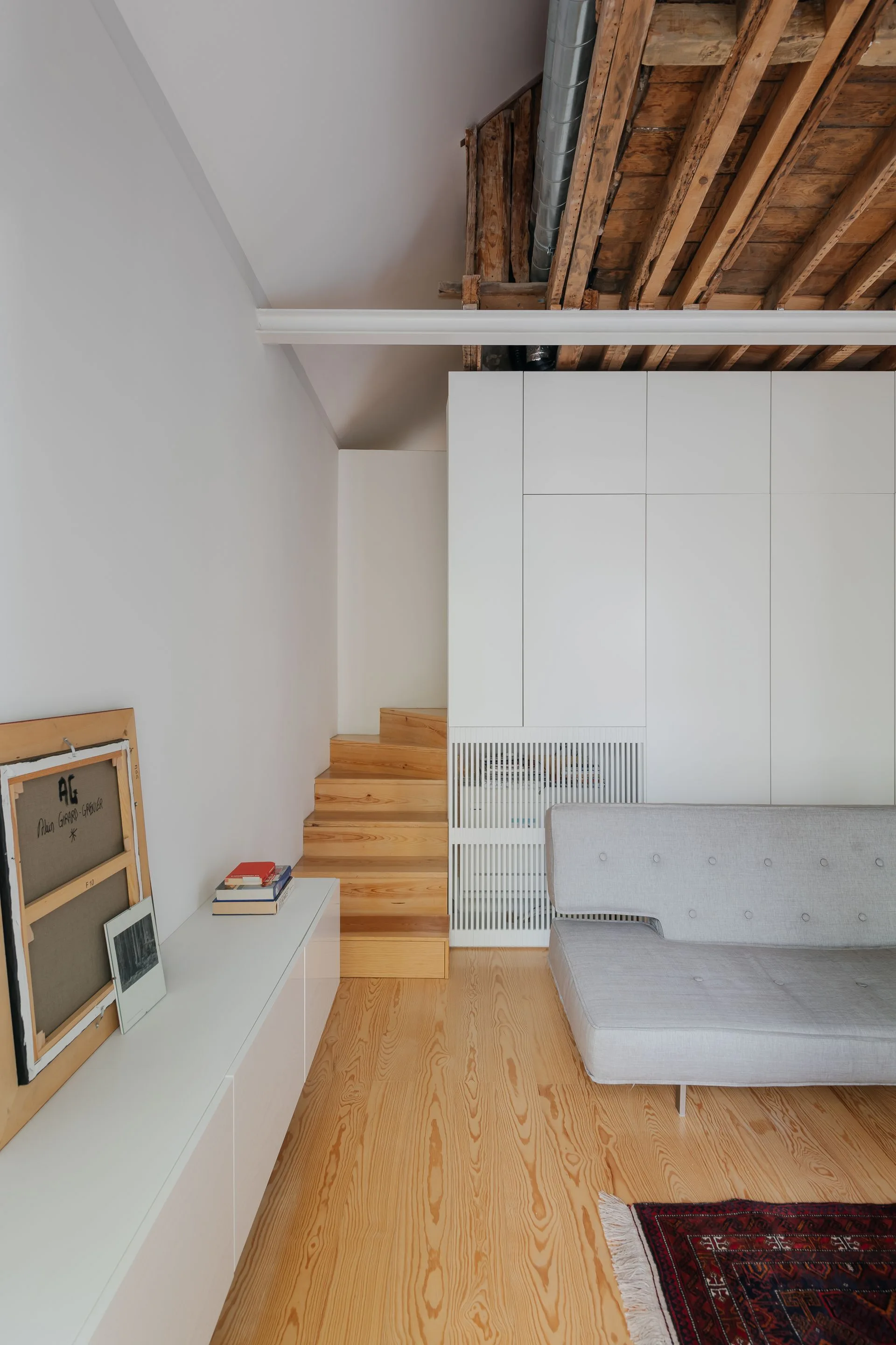 A minimalist living room features a grey sofa, light wood floors, a white built-in storage unit, and an exposed raw wood ceiling.