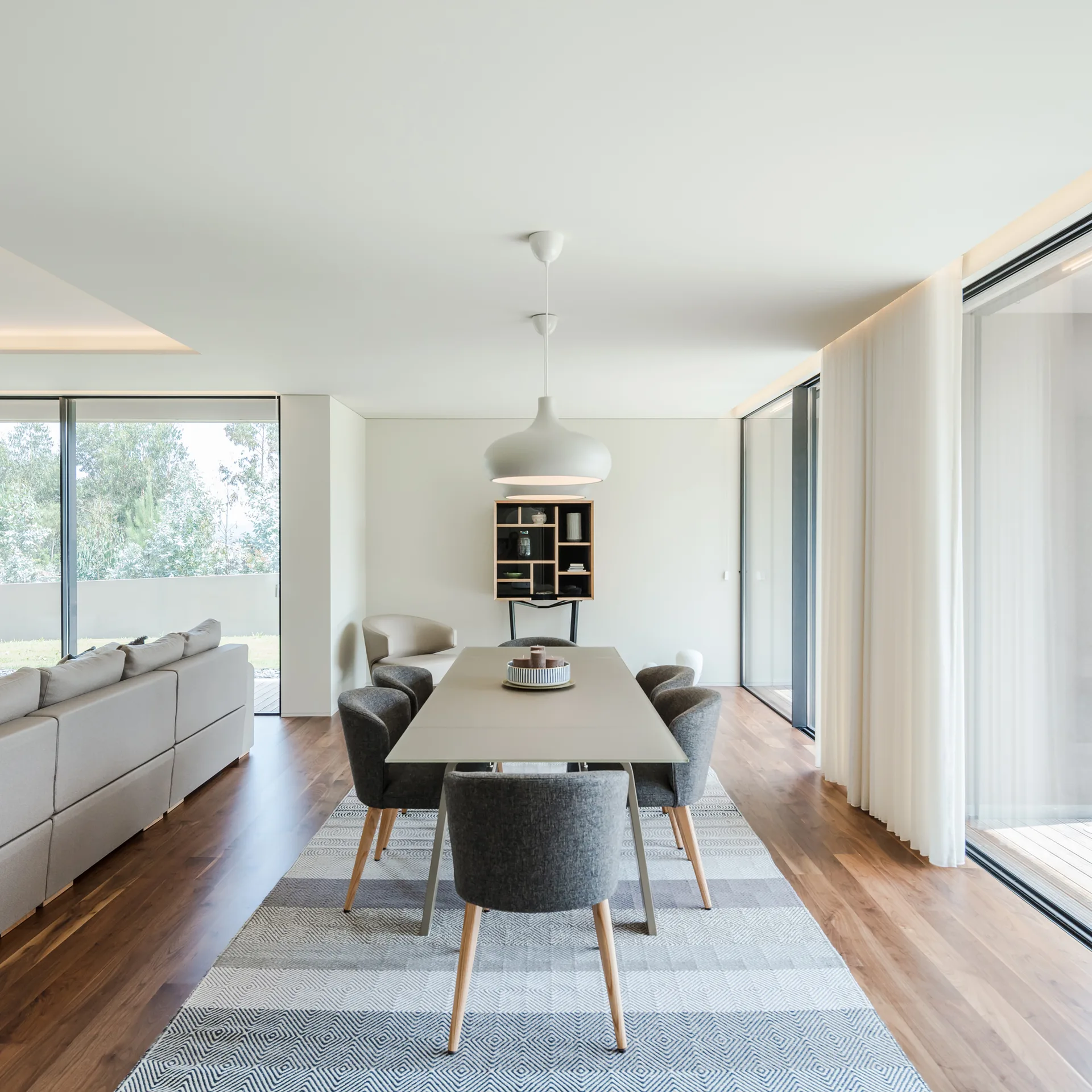A minimalist dining room features a grey table and chairs on a geometric rug, with floor-to-ceiling glass doors and a white pendant light.