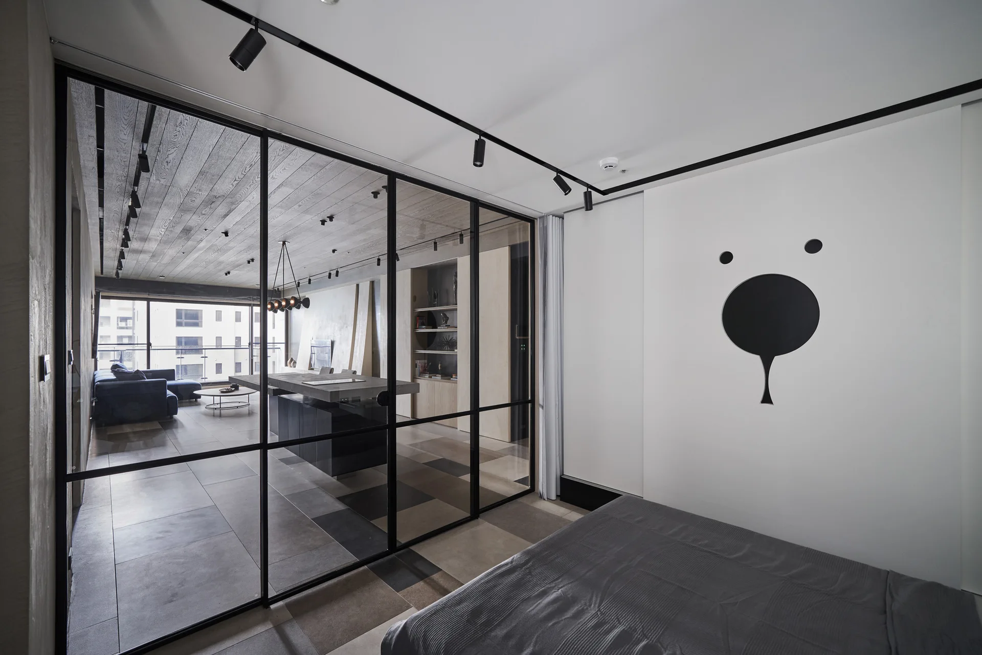 A modern grey bed is separated from an open living space by a black-framed glass partition wall and black track lighting.