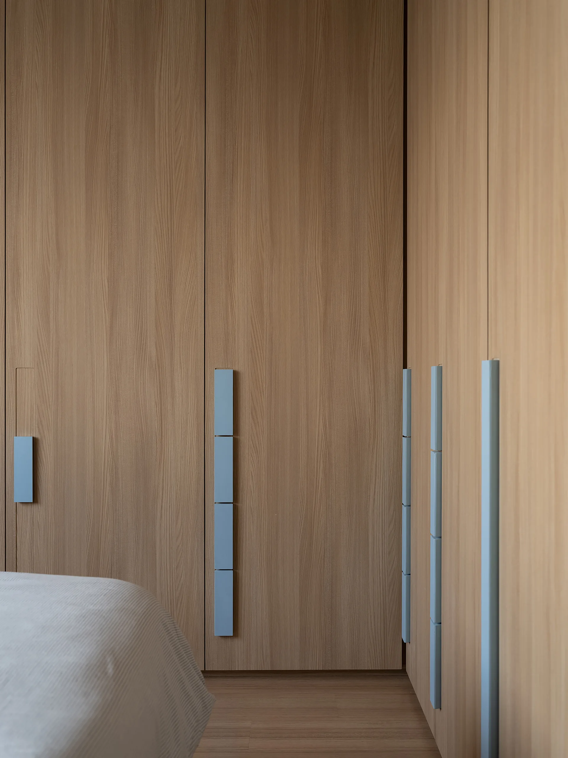 Minimalist light wood wardrobe doors with long, vertical blue block handles stand next to a partially visible bed on wood flooring.