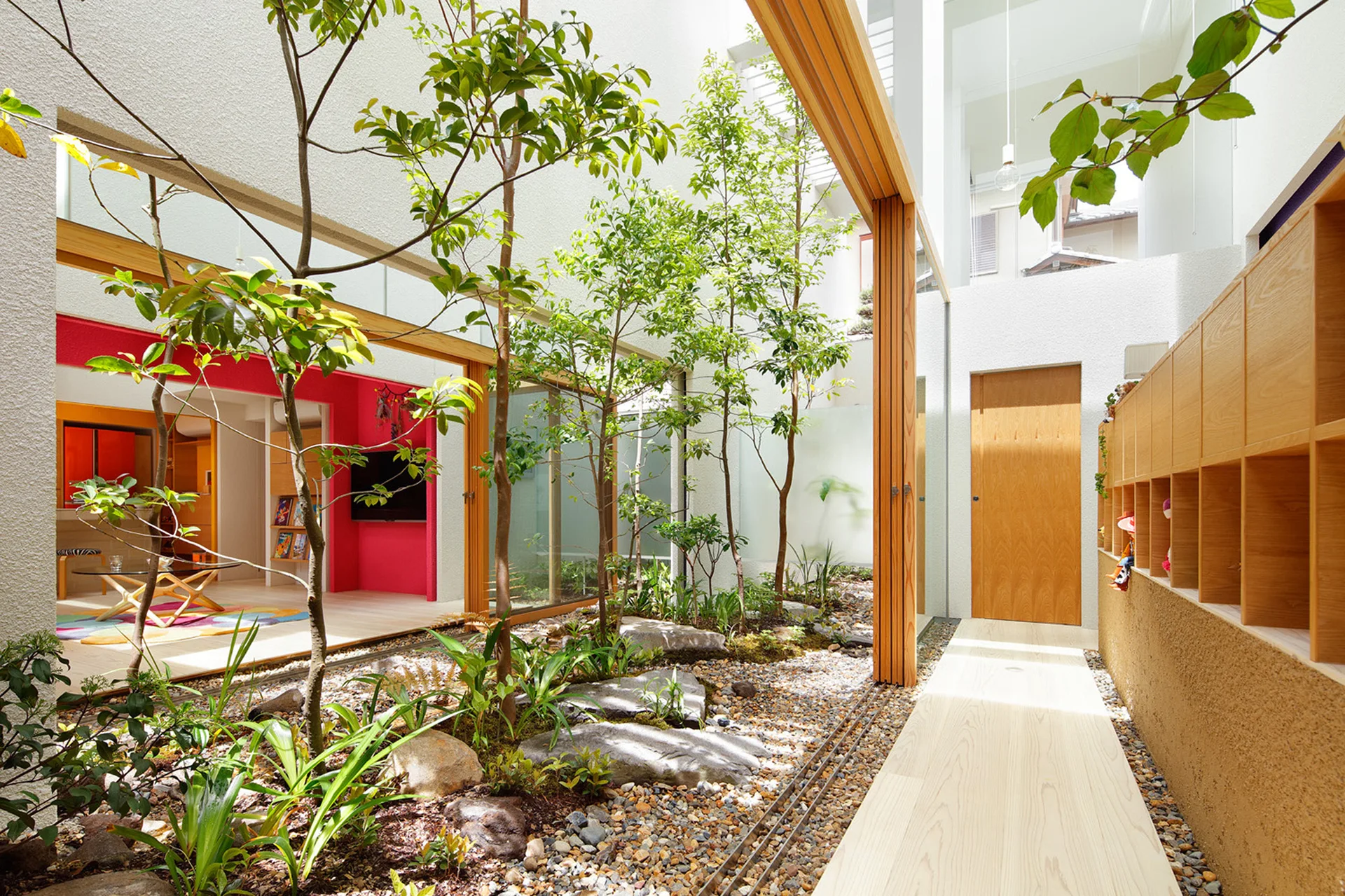 A modern residential courtyard features green trees, gravel, and large stones bordered by sliding wood-framed glass doors.