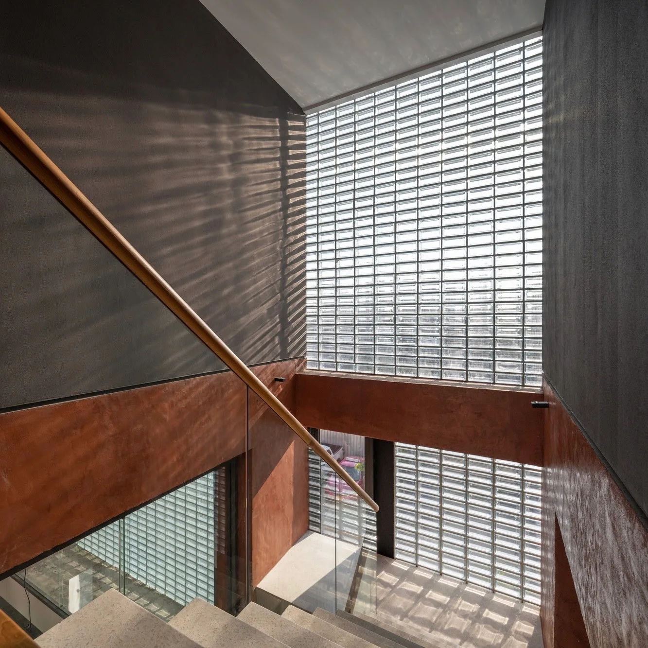 Looking down a concrete staircase with a wooden handrail, flanked by dark grey and terracotta walls, next to a large glass block wall.