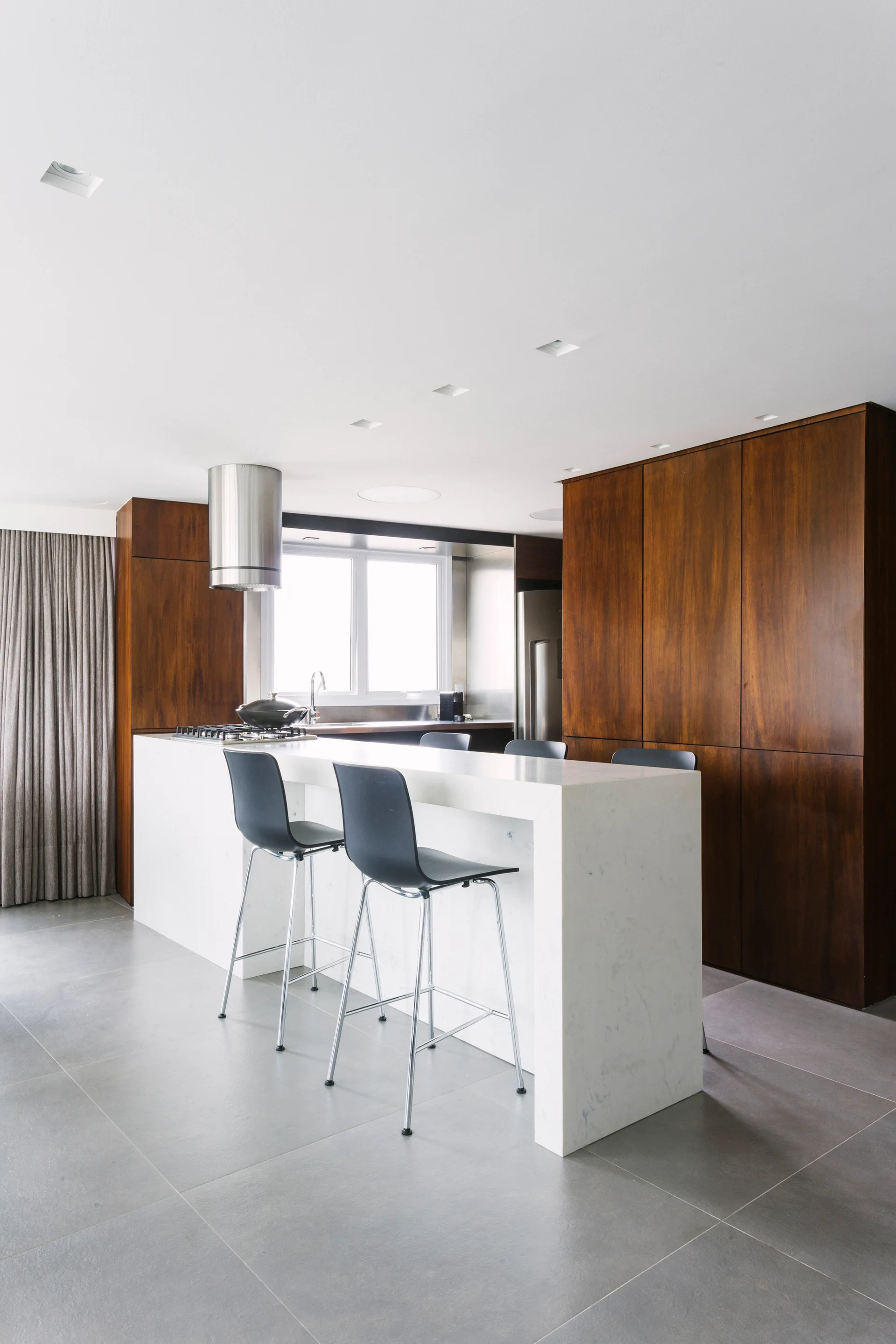 A modern kitchen featuring a white stone waterfall island with black bar stools, dark wood cabinets, and large grey floor tiles.