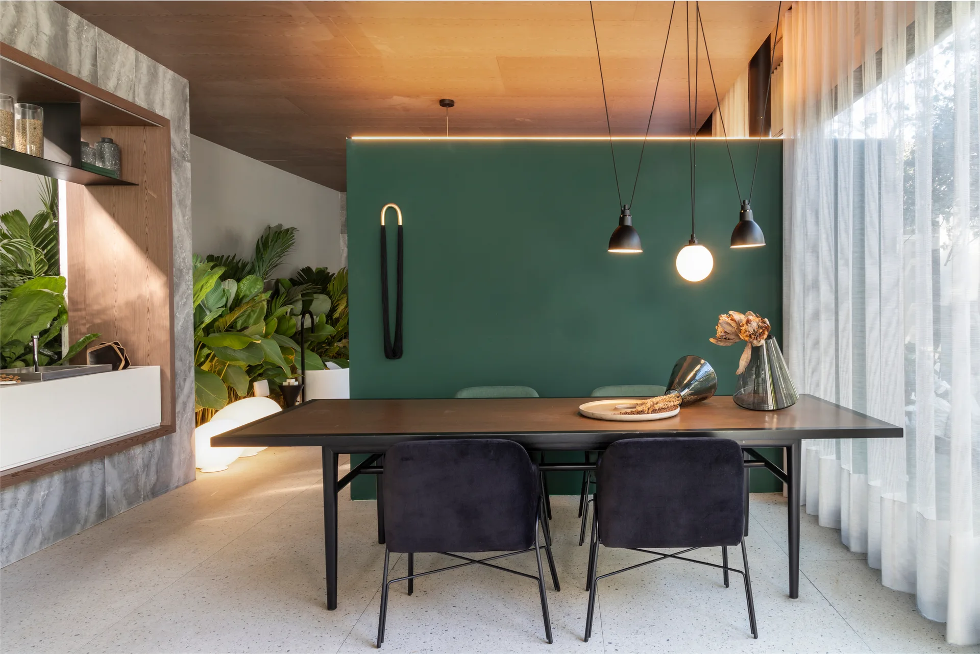 A contemporary dining room features a dark wood table with dark velvet chairs positioned in front of a forest green divider wall beneath a wood ceiling.