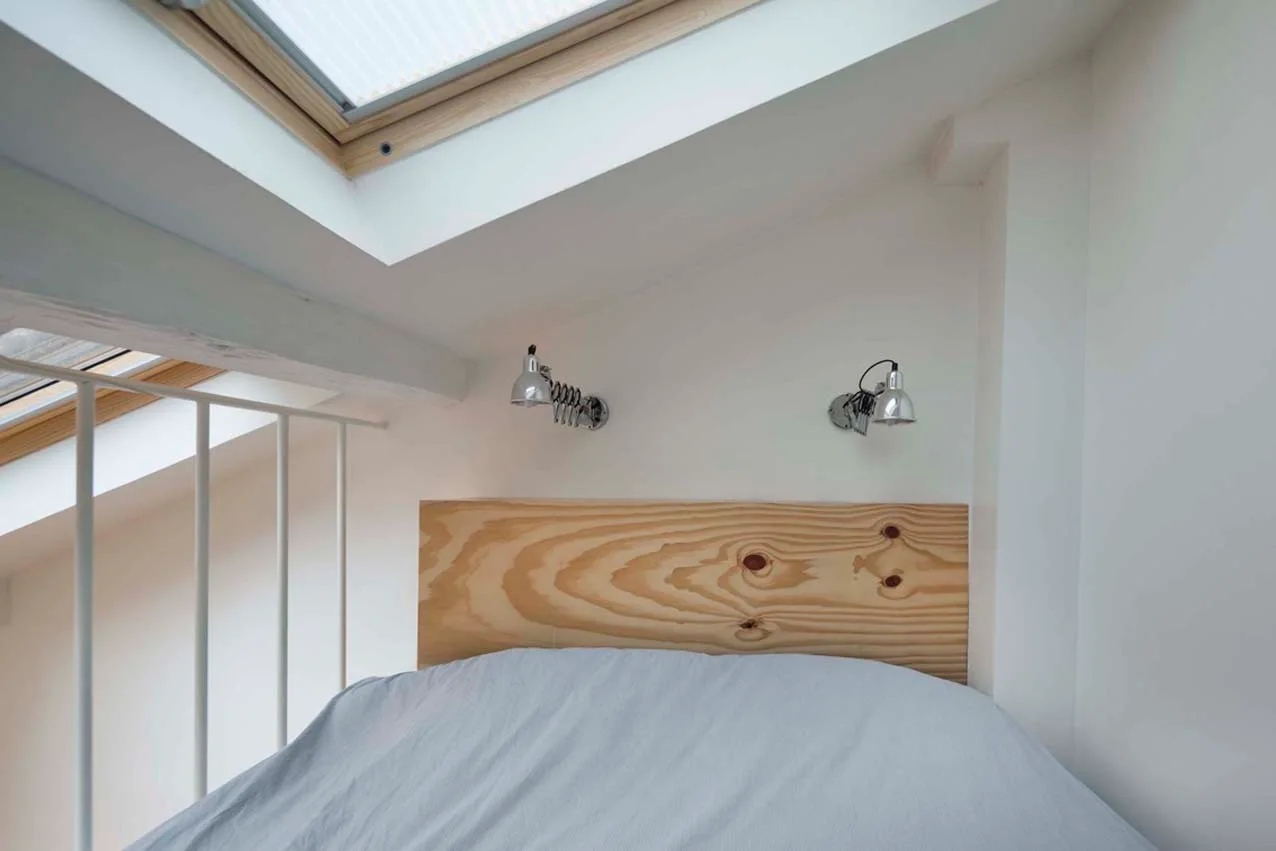 An attic loft bed with pale bedding features a raw pine wood headboard beneath a sloped white ceiling with a skylight.