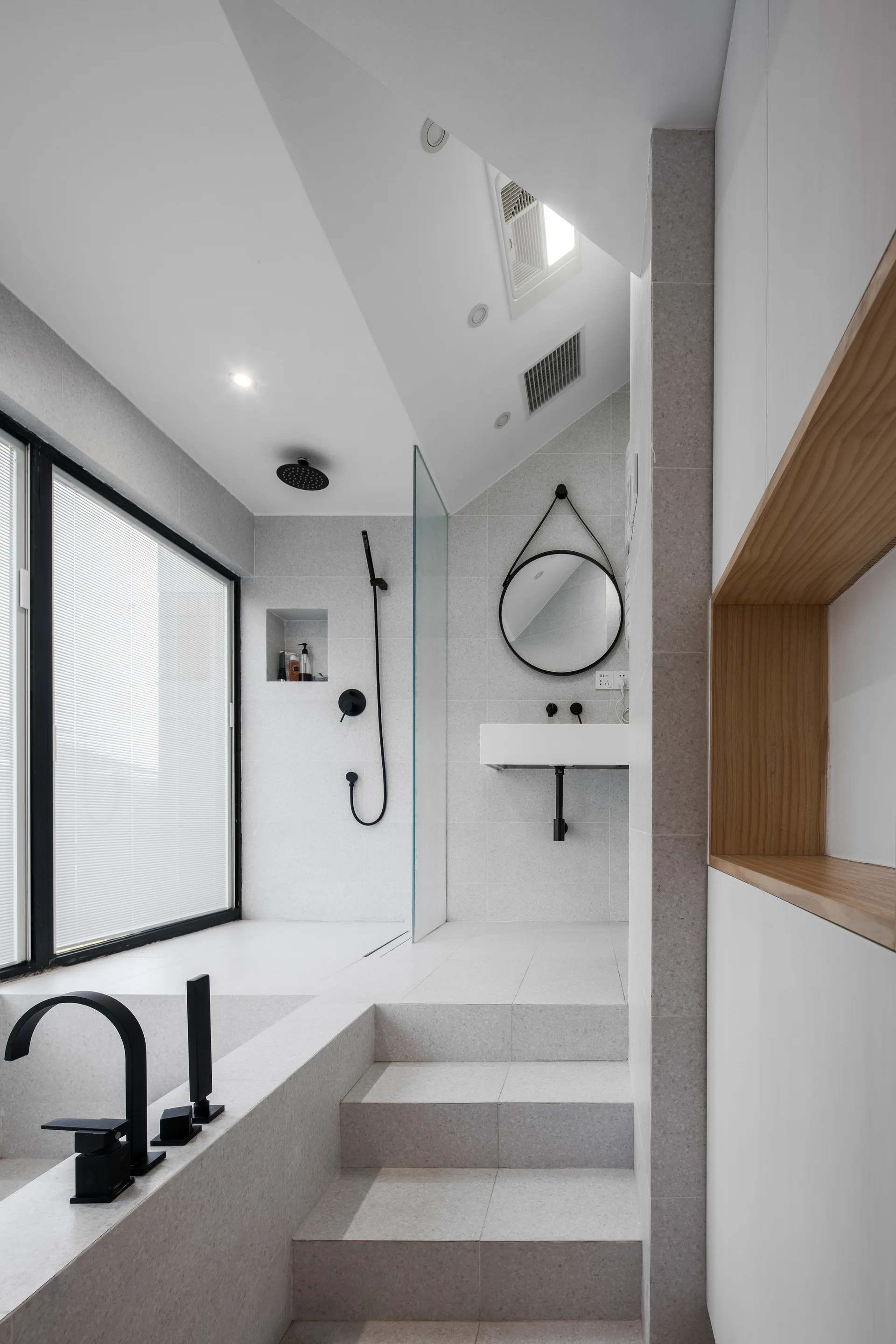 A modern bathroom with light gray tiled steps leading to a raised shower and bathtub area featuring matte black fixtures and a round mirror.