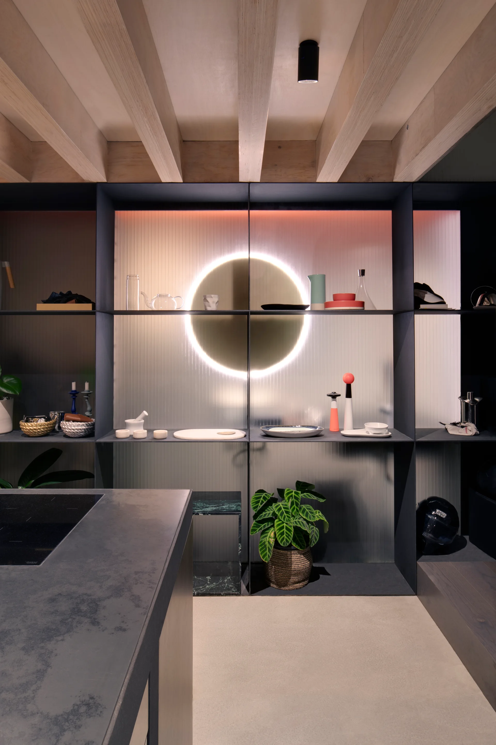 A dark metal open shelving unit in a kitchen features a circular backlight, ribbed wall panels, and exposed wooden ceiling beams.