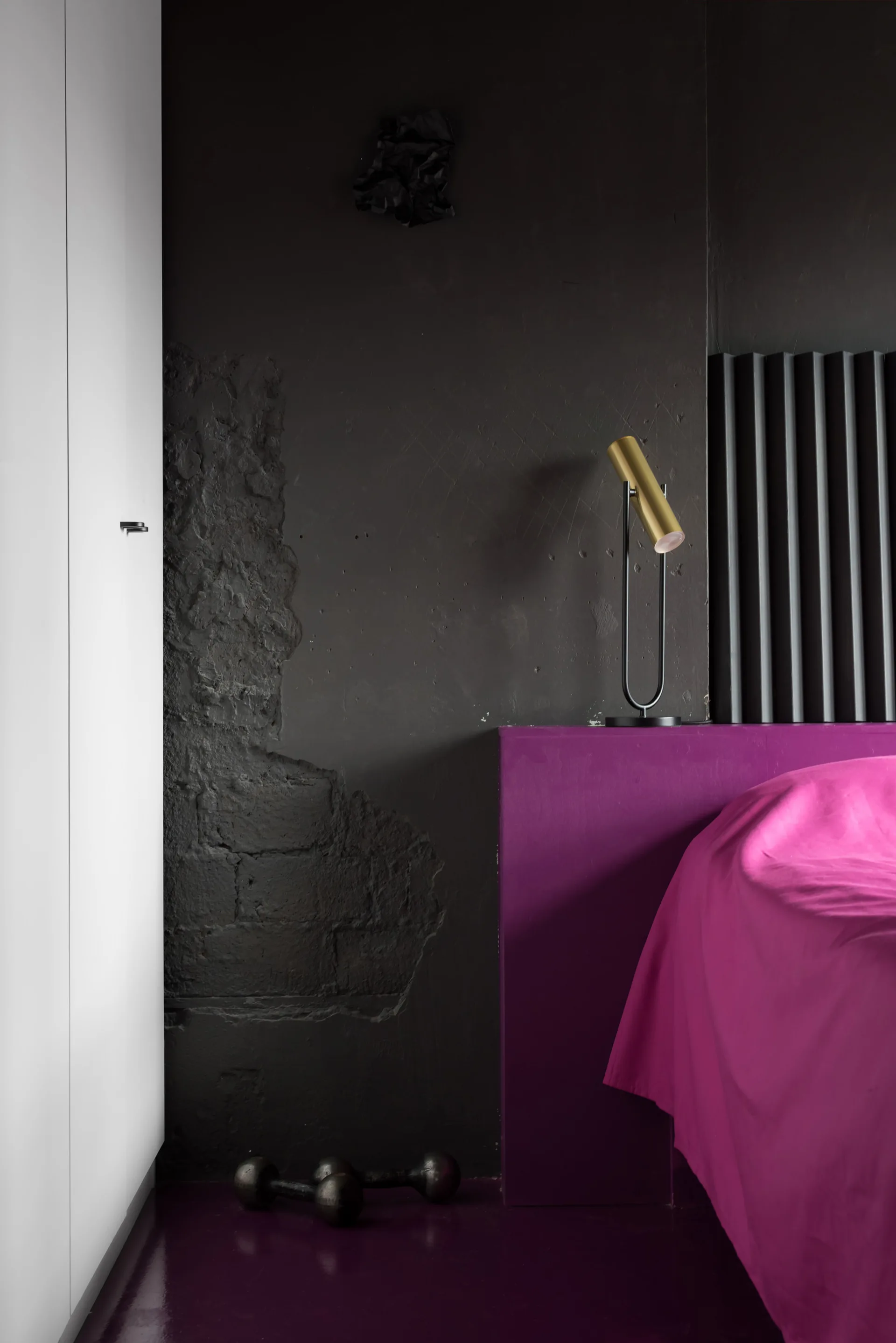 A bedroom corner showcases a textured black plaster wall, a sleek white wardrobe, and a brass cylindrical lamp resting on a magenta platform bed.