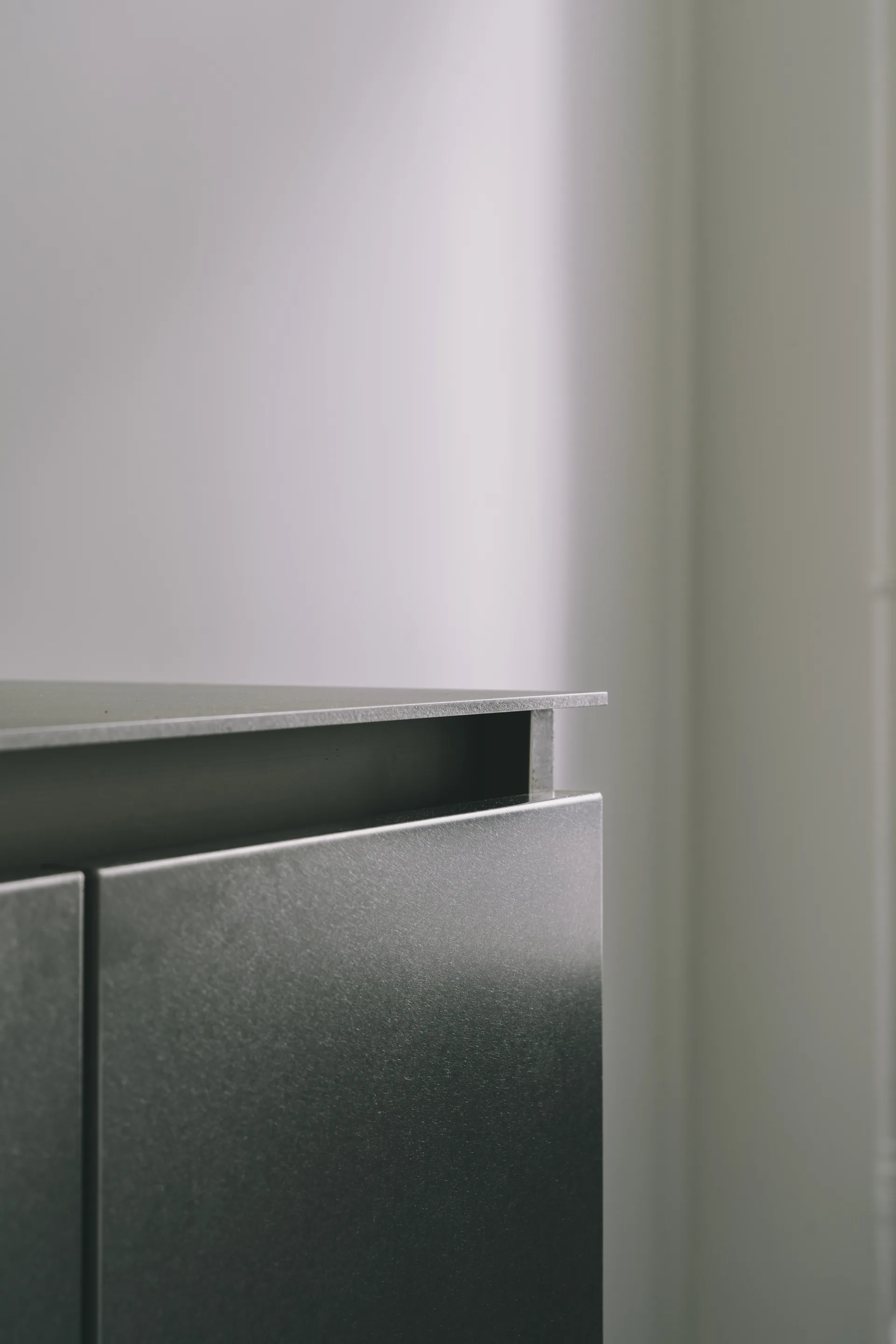 A close-up shot of a grey brushed metal cabinet featuring a recessed top channel and sharp geometric edges against a plain light wall.