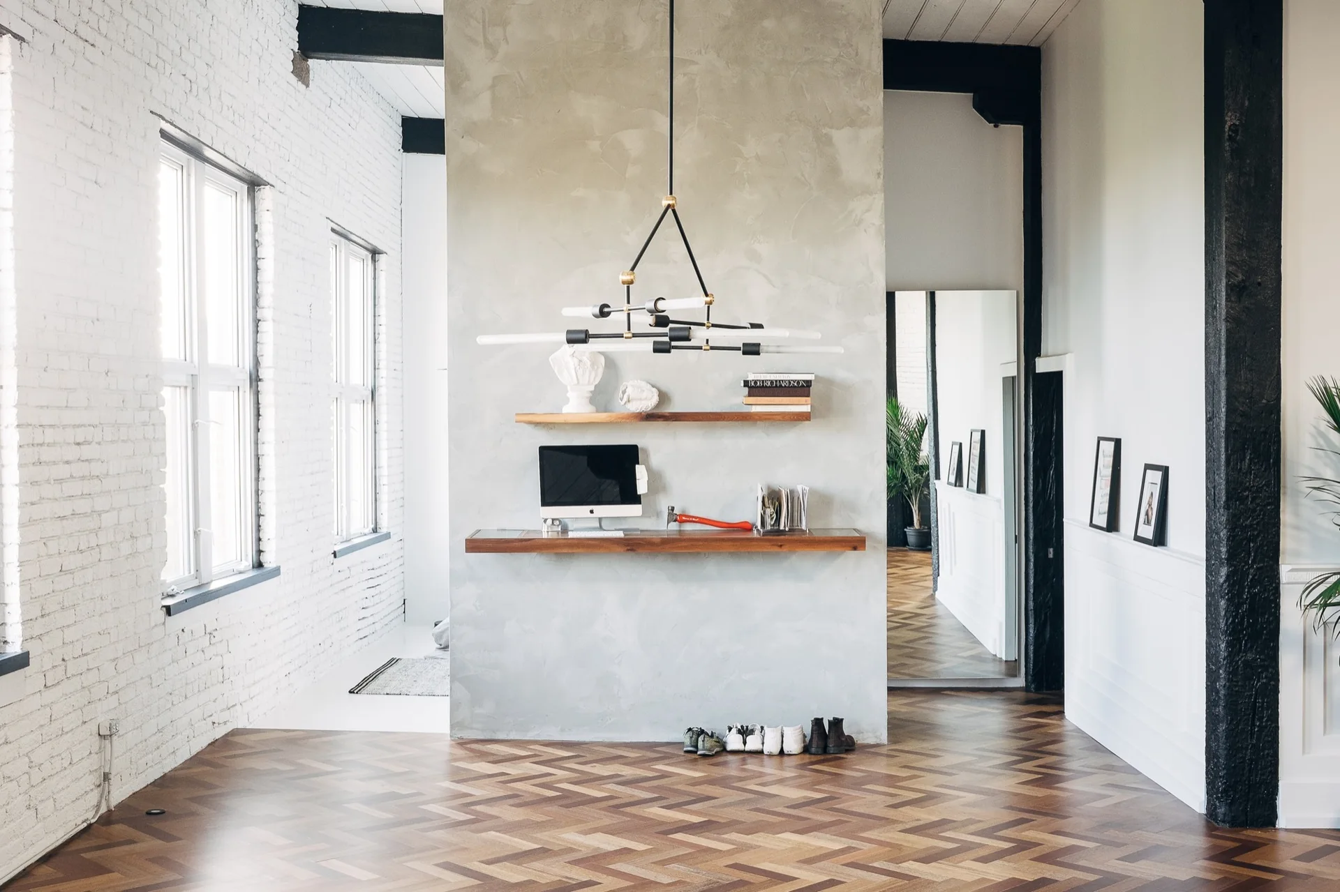 A home office features a floating wood desk on a grey plaster partition wall, flanked by white brick walls and herringbone flooring.