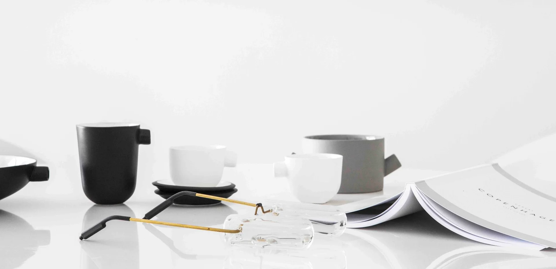 A minimalist arrangement of matte ceramic cups, an open book, and thin metal glasses on a reflective white surface.