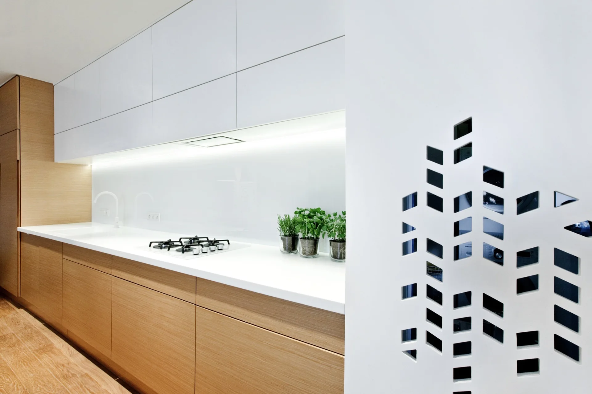 A modern minimalist kitchen features wood lower cabinets, white upper cabinets, an illuminated backsplash, and a white partition with geometric cutouts.