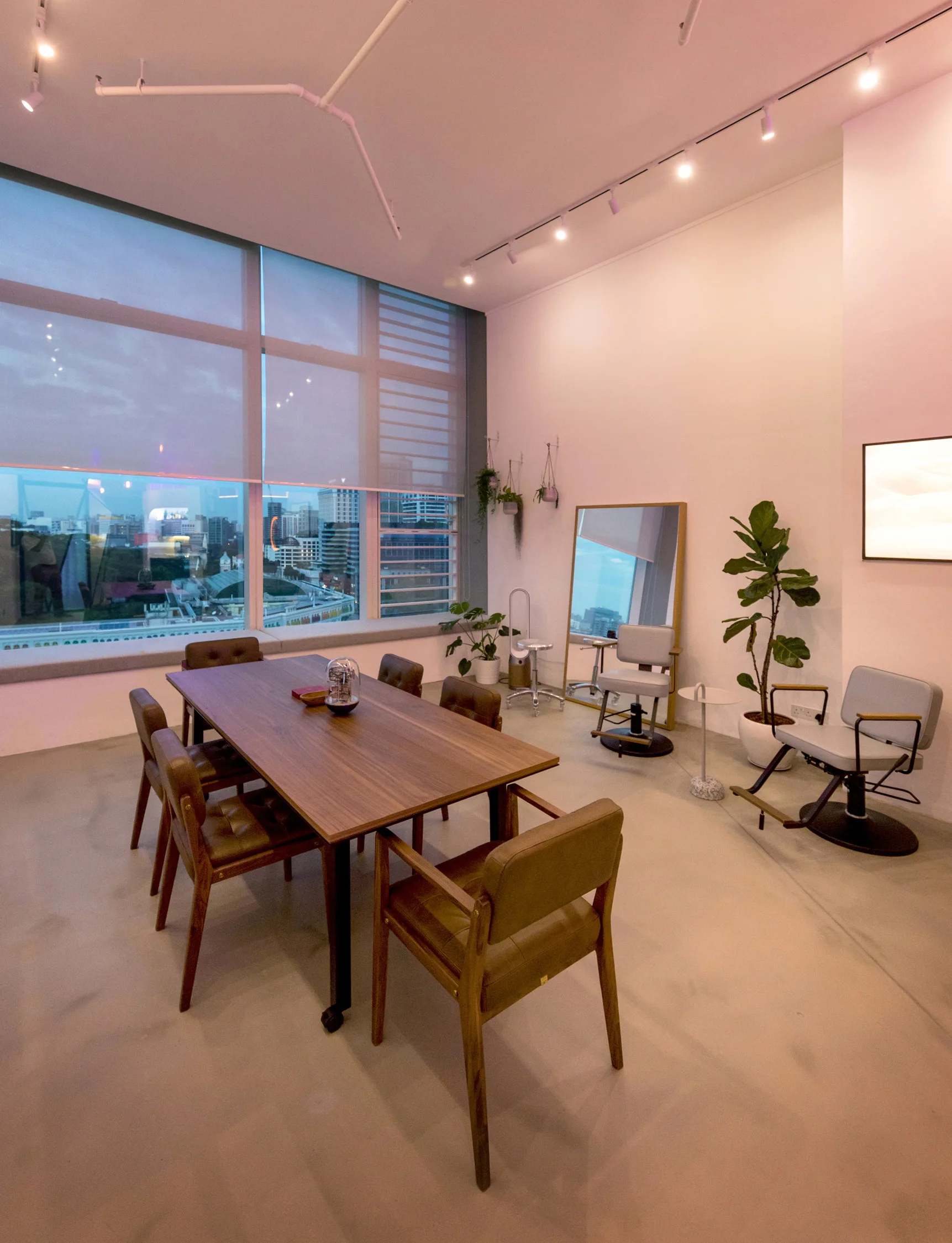 A modern multi-use room features a wooden dining table with chairs, two salon chairs, a floor mirror, and a large window overlooking the city.