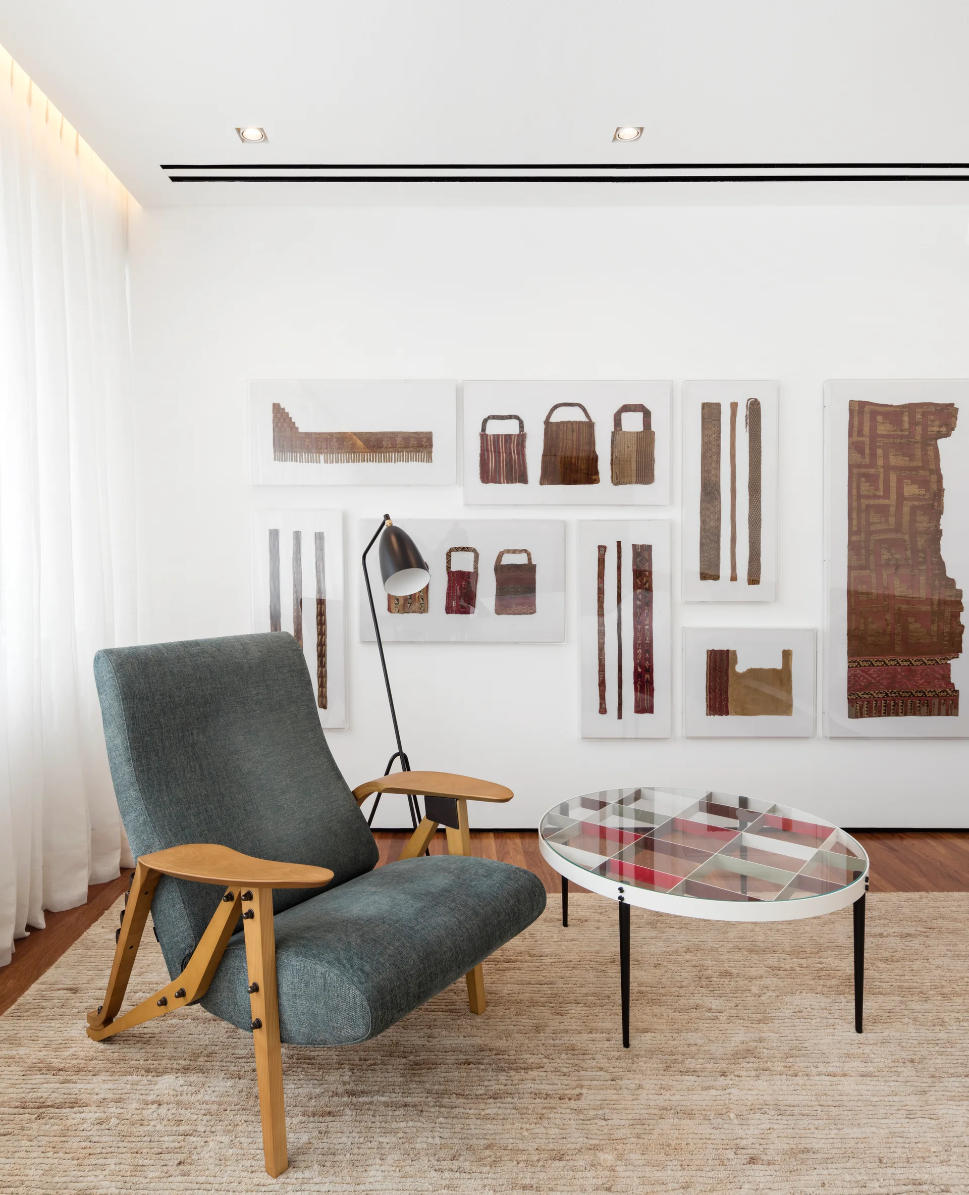 A mid-century blue armchair and oval glass coffee table sit on a beige rug near a white wall with framed textile art.