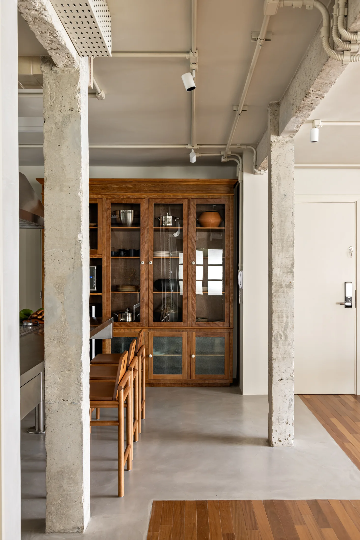 An industrial kitchen features raw concrete columns, a tall wooden china cabinet with glass doors, wooden bar stools, and track lighting.