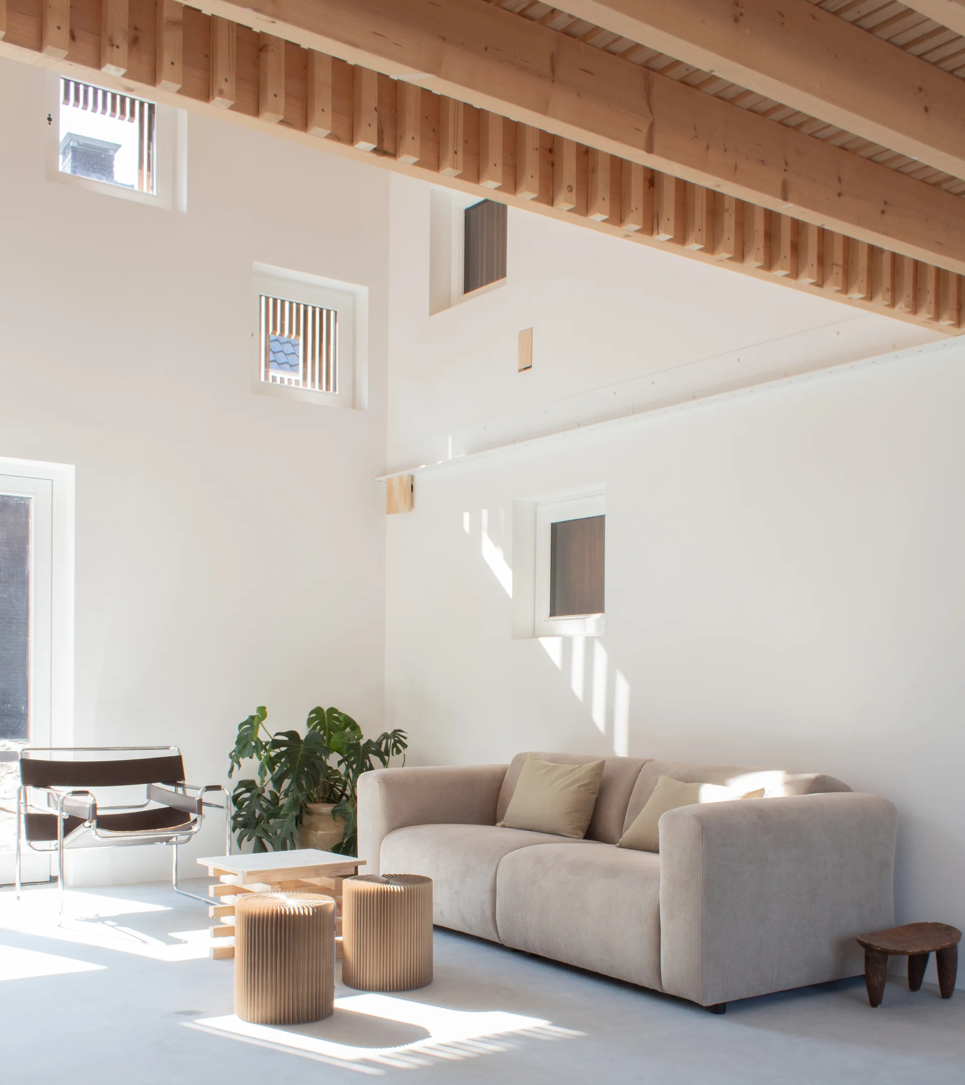A minimalist white living room features a neutral beige sofa, two ribbed cardboard side tables, and prominent exposed wooden ceiling beams.