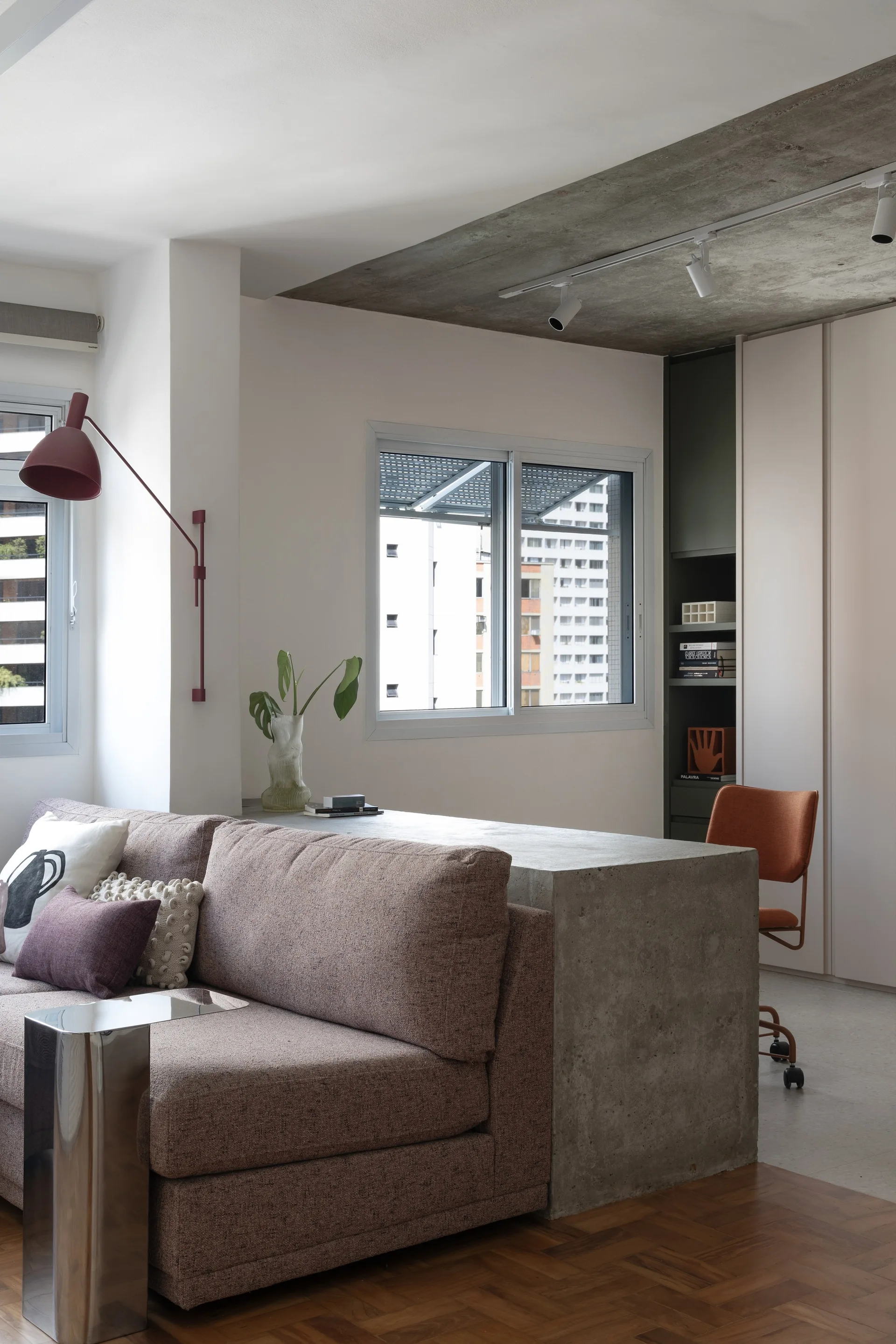 A modern living room featuring a mauve textured sofa backed by a concrete desk, an orange office chair, track lighting, and a concrete ceiling.