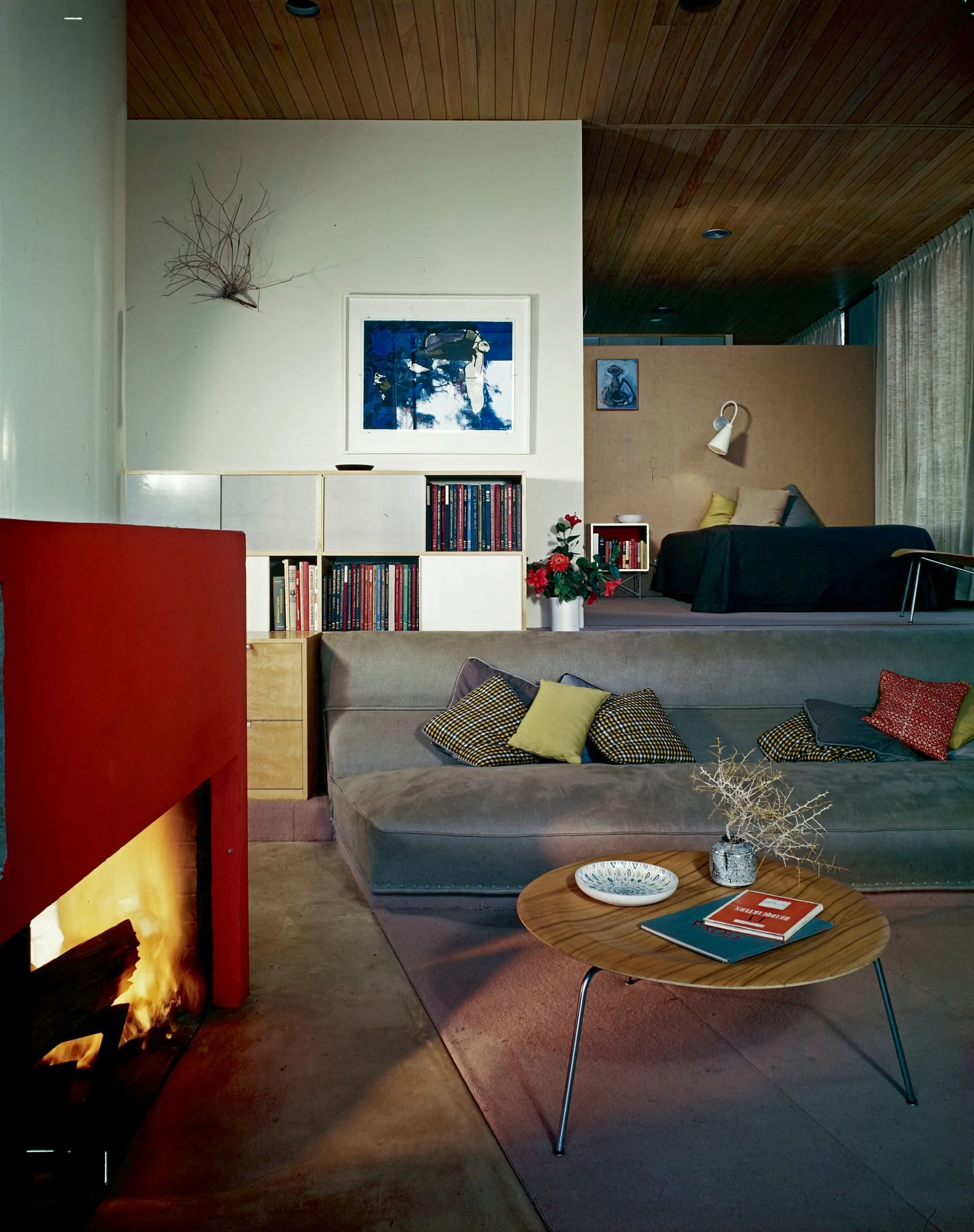 A mid-century modern split-level living room featuring a sunken gray sofa, a round wood coffee table, a bright red fireplace, and a raised bedroom.
