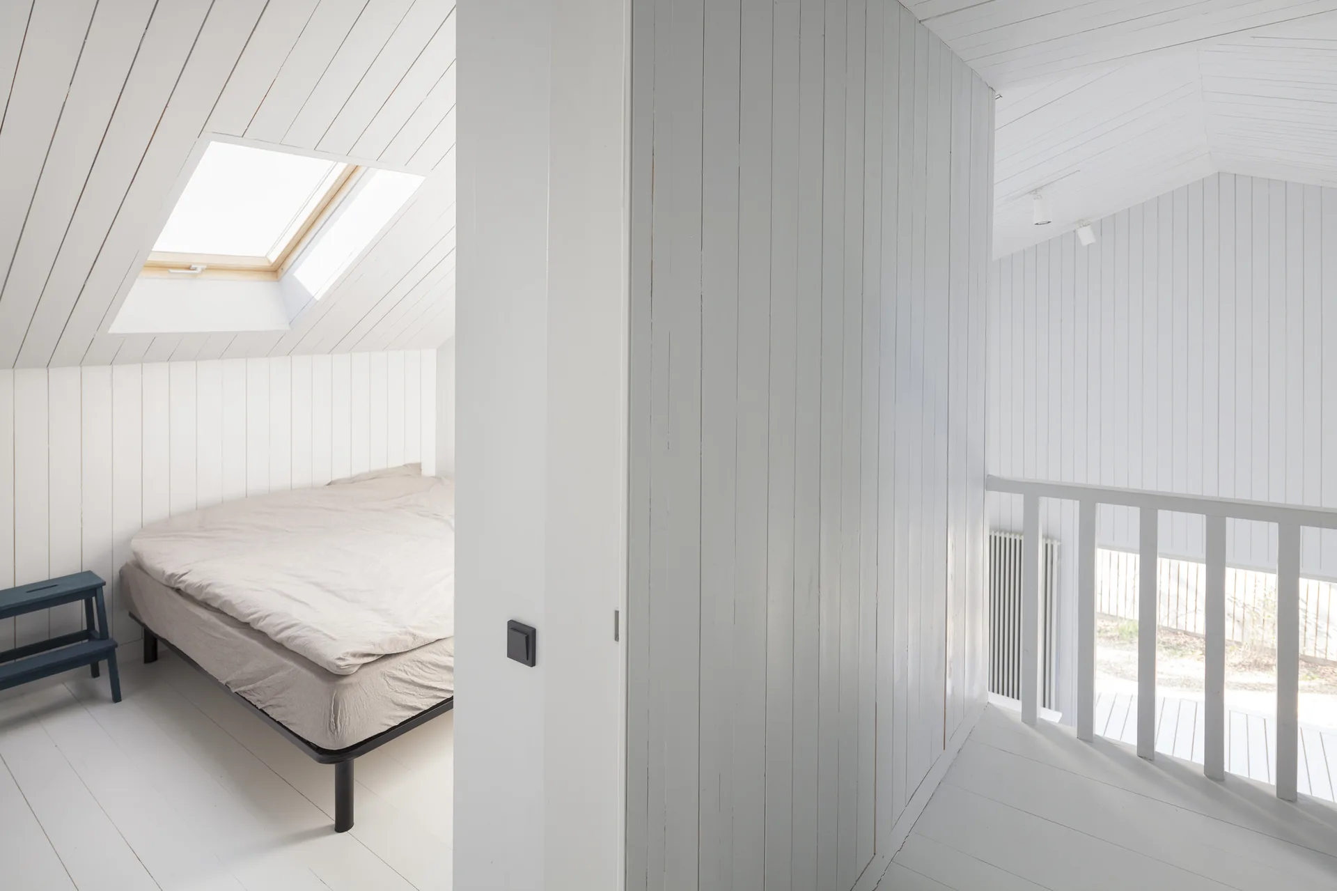 A minimalist attic bedroom featuring white vertical wood paneling on the walls and ceiling, a low bed under a skylight, and white painted floors.