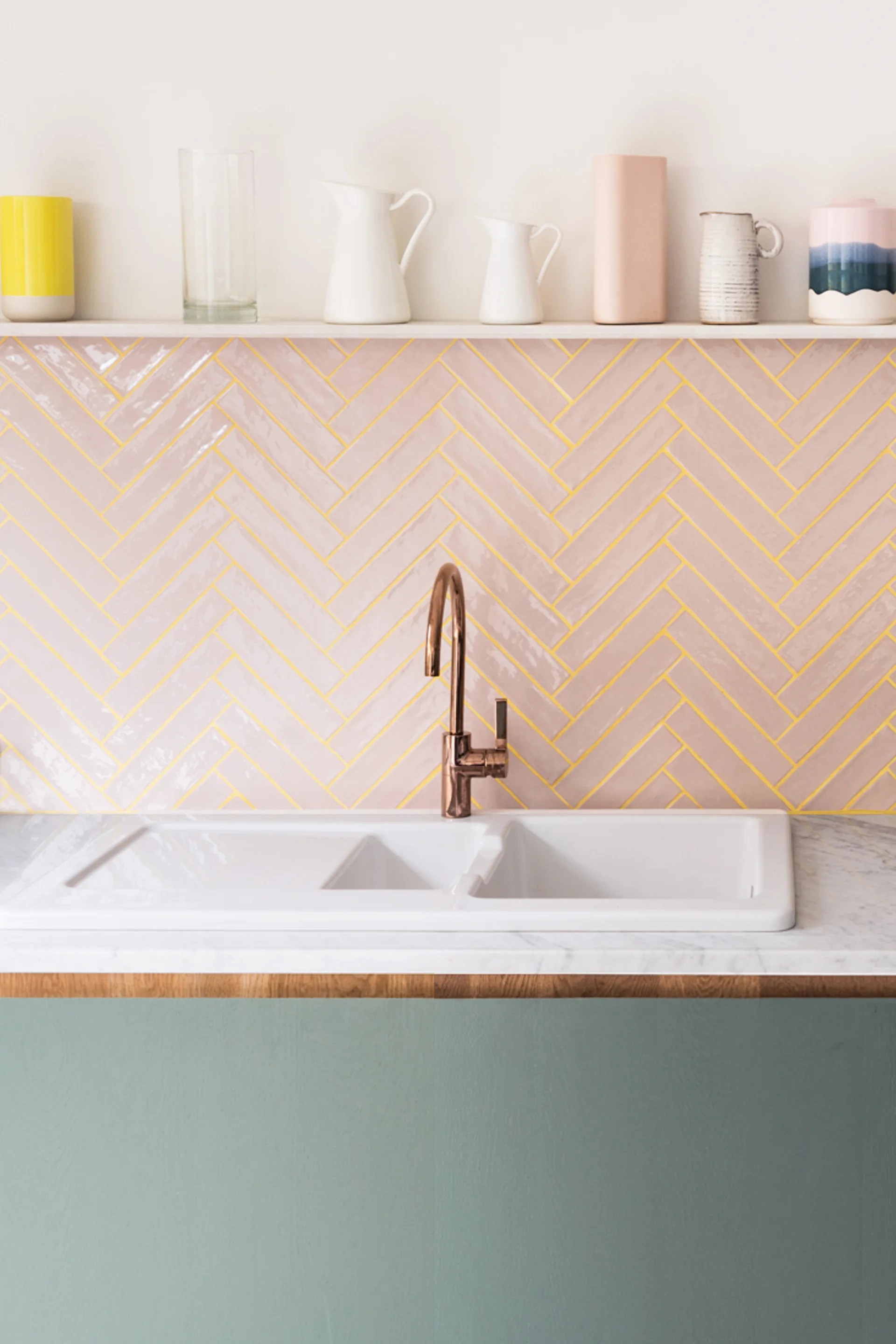 A white double sink with a copper faucet is set against a pink herringbone tile backsplash with yellow grout and a shelf holding vases.