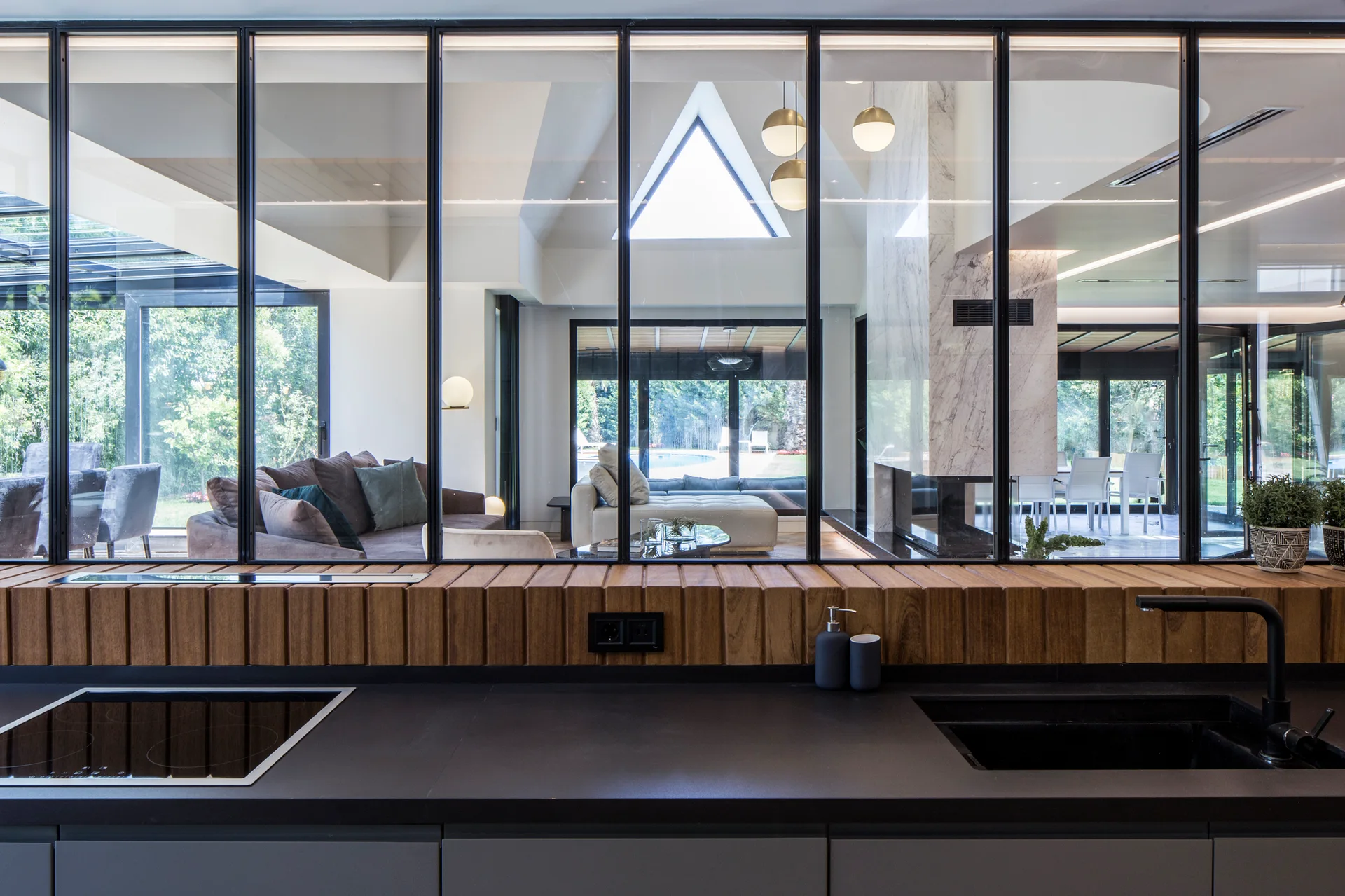 A modern kitchen countertop with a sink and stovetop sits before a black-framed glass partition looking into a spacious living room.