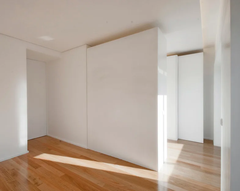 A minimalist white hallway features light-colored hardwood floors, a central white partition wall, and integrated flush cabinetry.