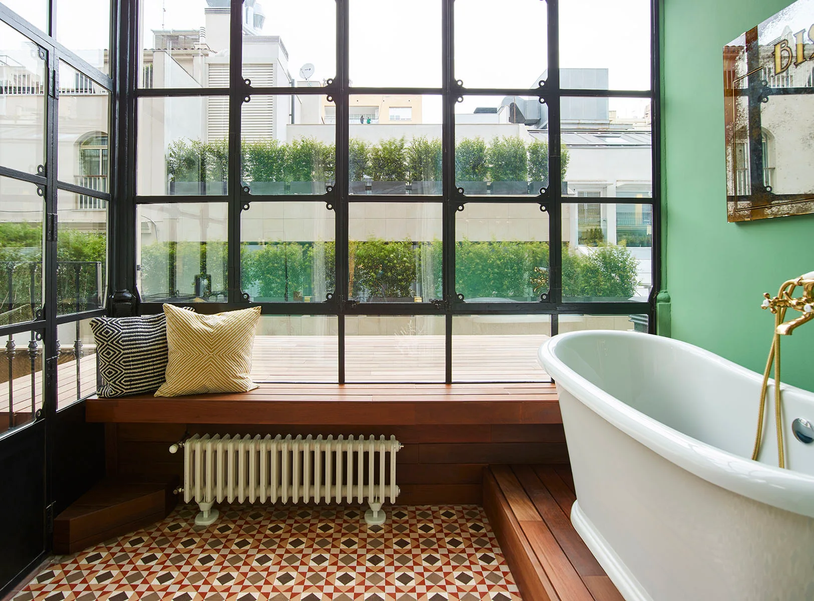 A white freestanding bathtub sits on a wooden platform next to a green wall, with patterned tile floors and large black grid windows.