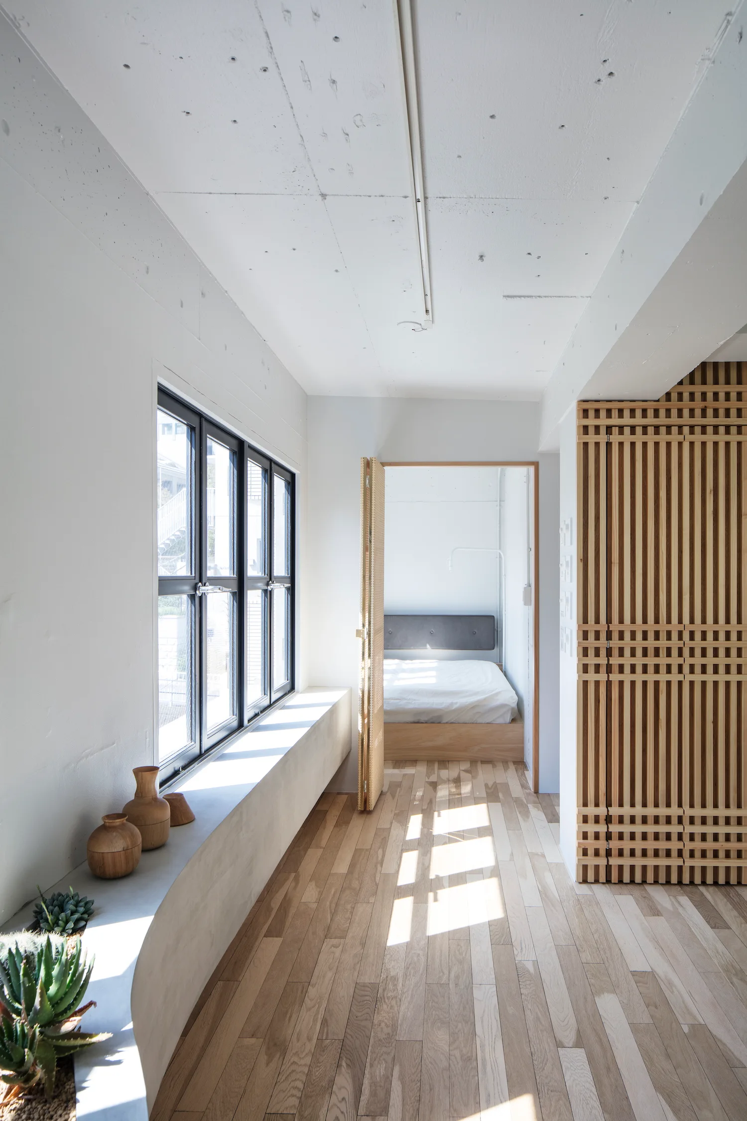 A minimalist hallway with light wood floors, a long concrete window ledge, and a tall louvered wooden closet leading to a platform bed.