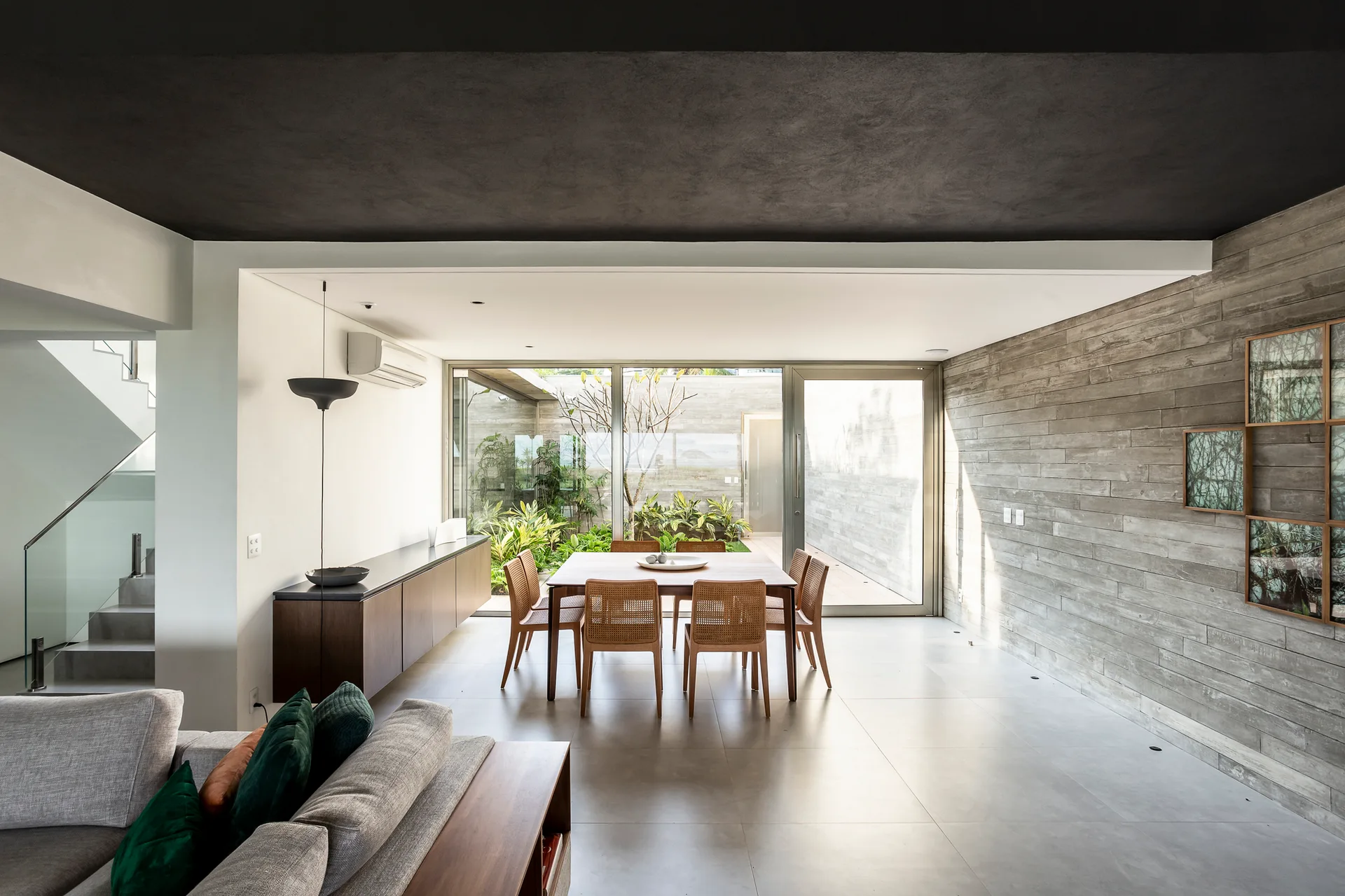 A modern dining room features a wooden table and woven chairs next to a large sliding glass door that opens to a green courtyard garden.