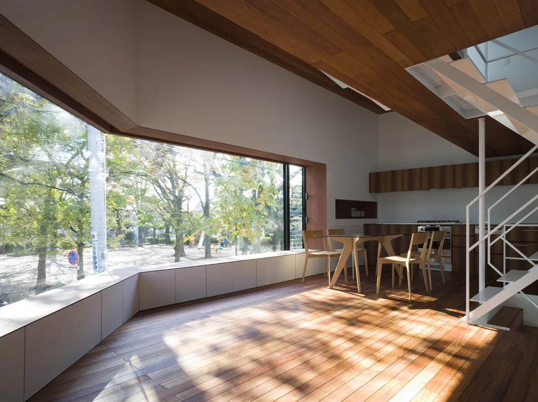A modern dining room with wood flooring, a white metal staircase, and a large panoramic window overlooking trees in a park.
