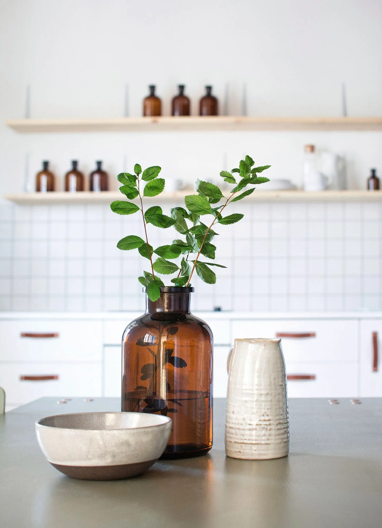 A close-up of a kitchen table with an amber glass vase, ceramic pitcher, and bowl in front of a white tiled backsplash and floating shelves.