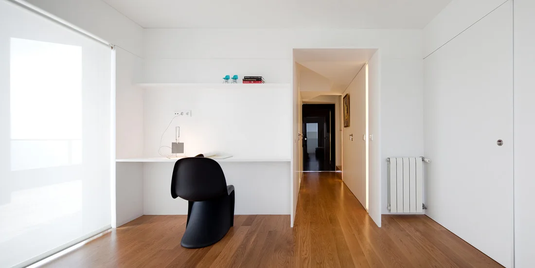 A minimalist white home office features a built-in desk, a sculptural black chair, wood flooring, and a floating shelf against a solid white wall.