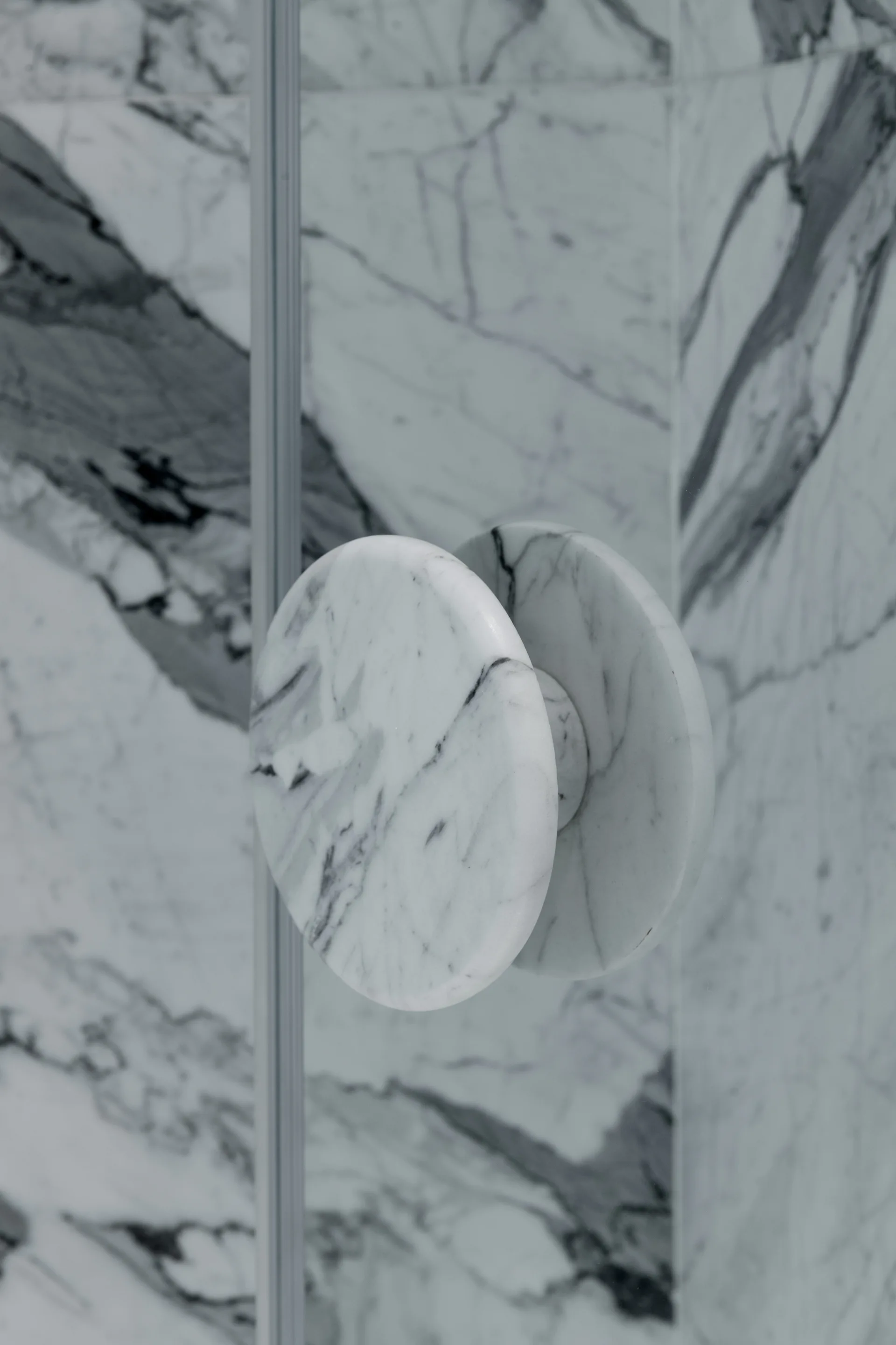 A close-up of a circular white marble handle on a glass shower door, with heavily veined marble wall tiles visible in the background.