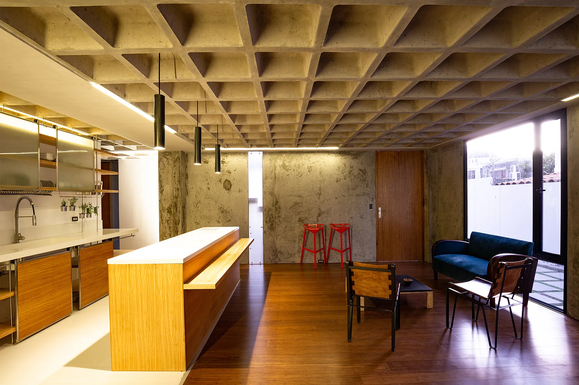 An open-concept kitchen and living area features an exposed concrete waffle ceiling, wooden flooring, a wooden island, and a blue sofa.