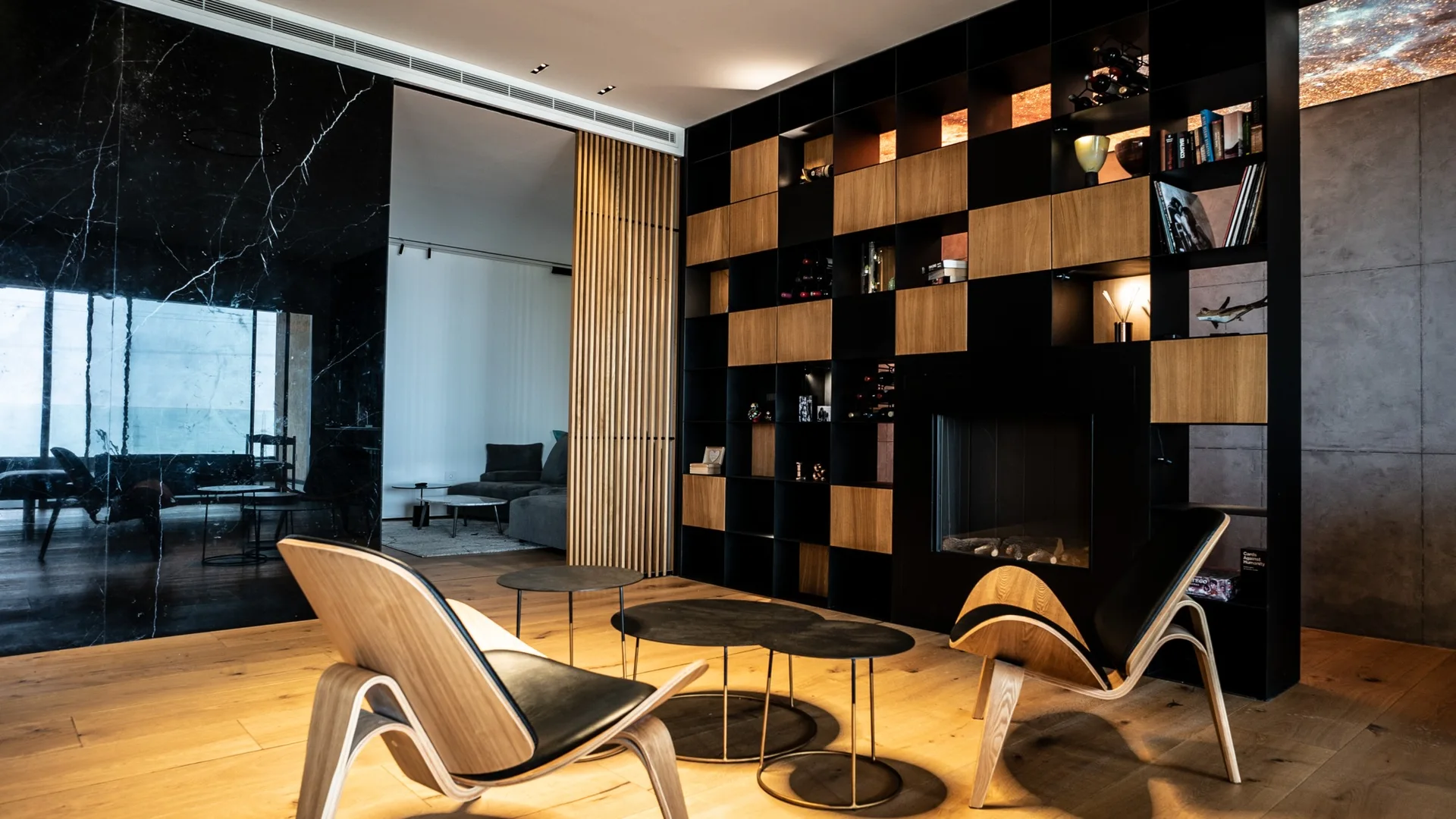 A modern lounge features two curved wooden chairs, small black coffee tables, a black and wood grid shelving unit with a fireplace, and a marble wall.