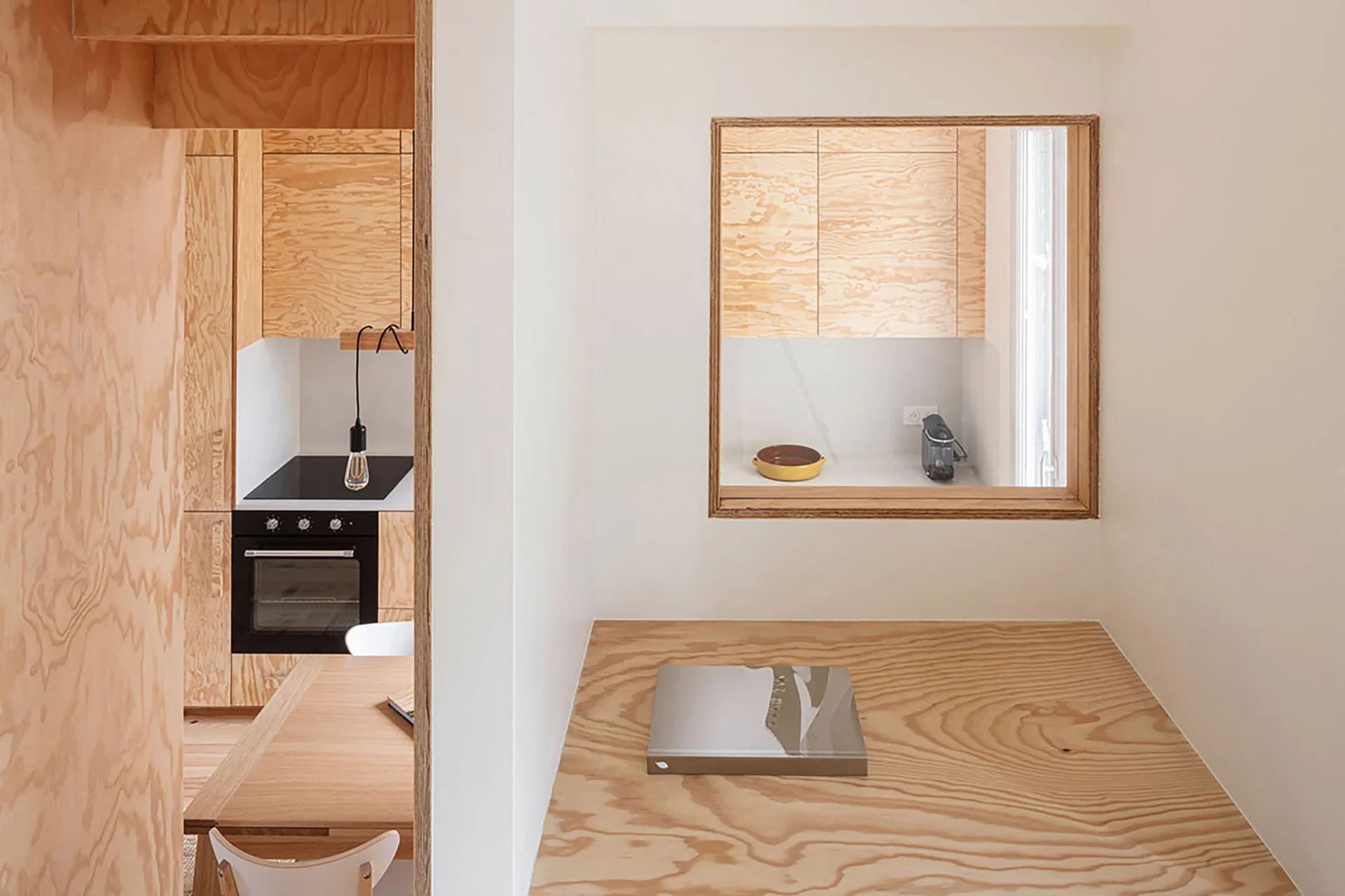 A minimalist kitchen featuring wood-grain plywood cabinetry, a black built-in oven, and a square pass-through window looking from a raised platform.