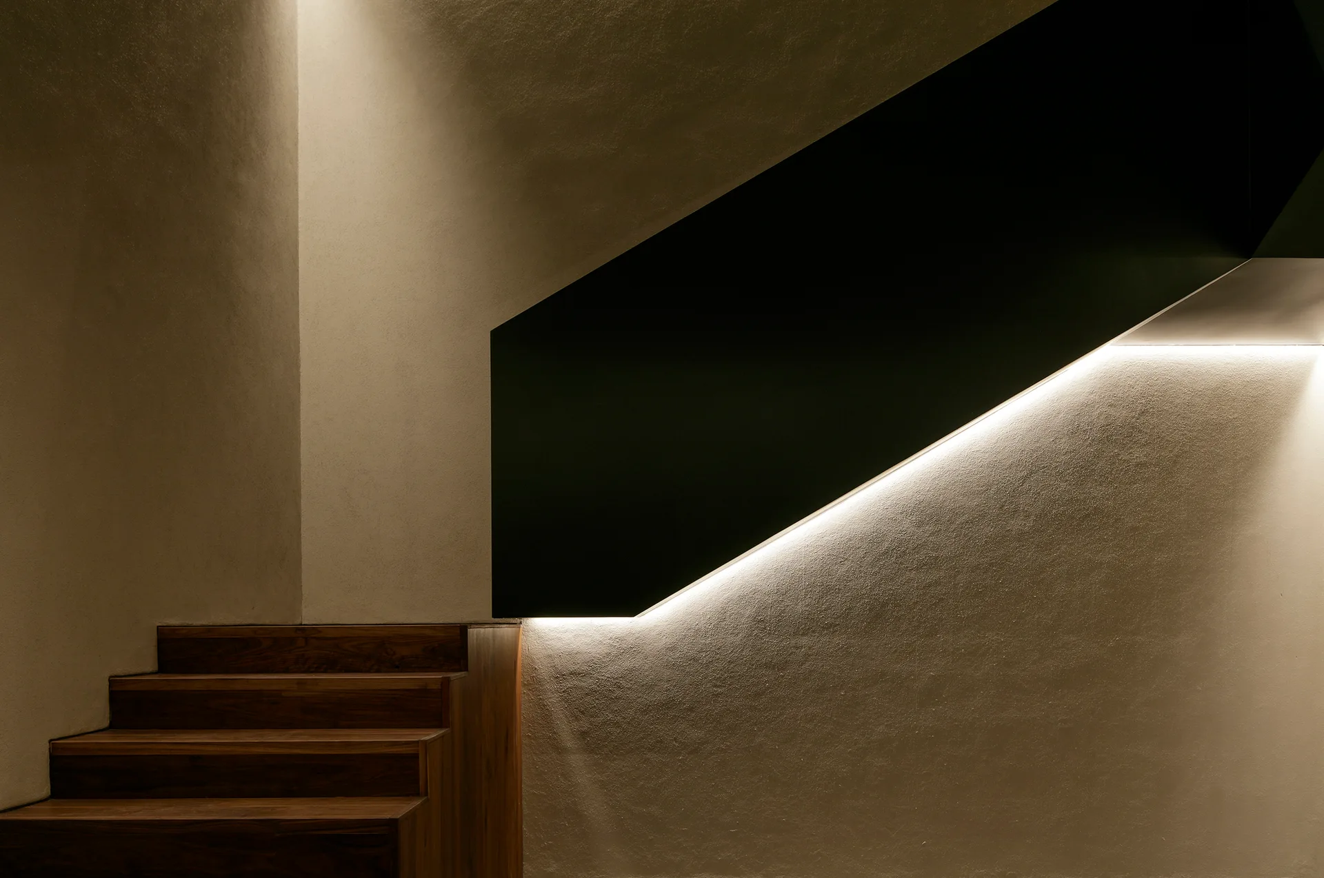A modern wooden staircase with a textured plaster wall and a solid black panel railing illuminated by a warm integrated LED light strip.
