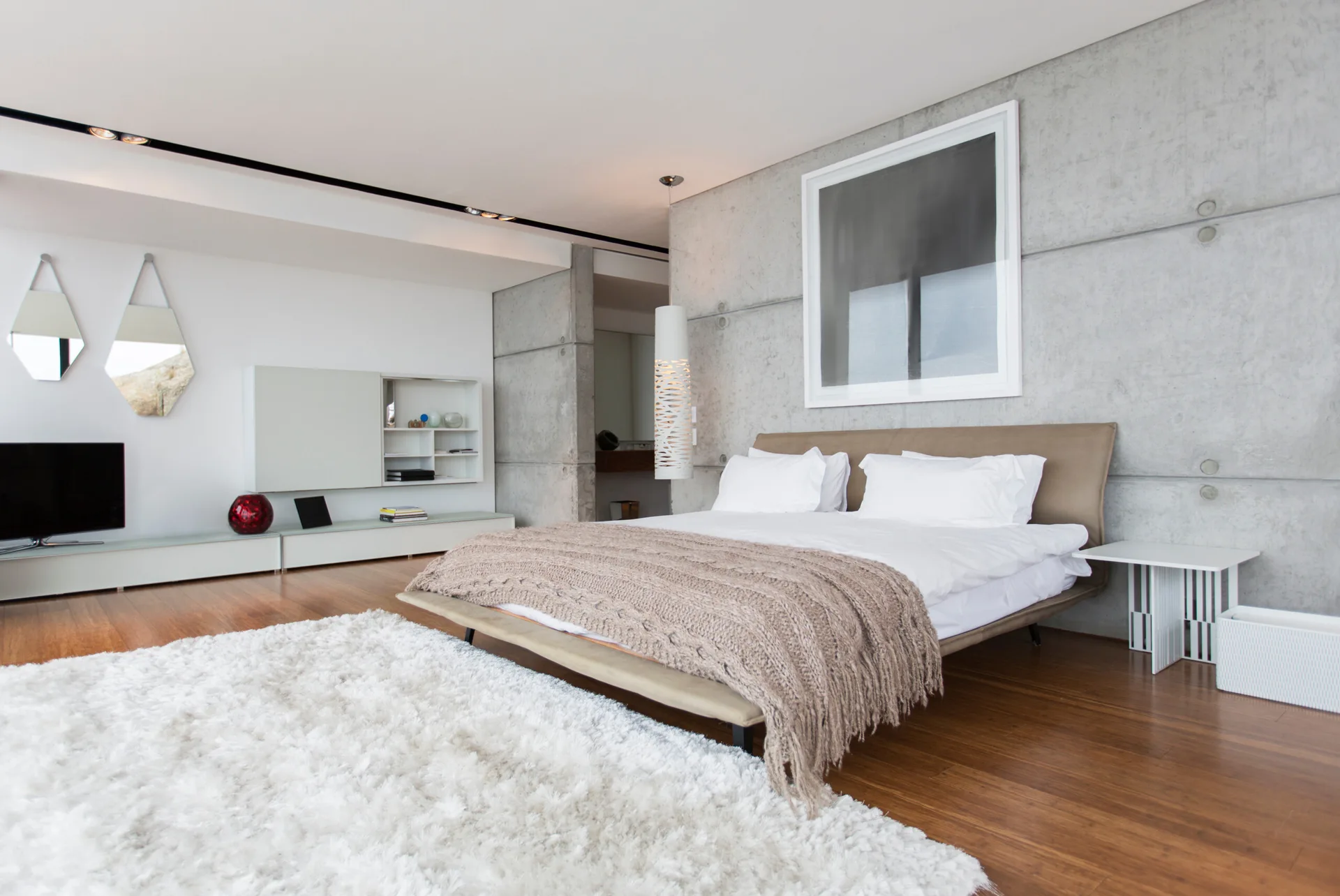 A contemporary bedroom features an exposed concrete panel wall, a tan upholstered bed with a knit throw, and a white shag rug on hardwood floors.