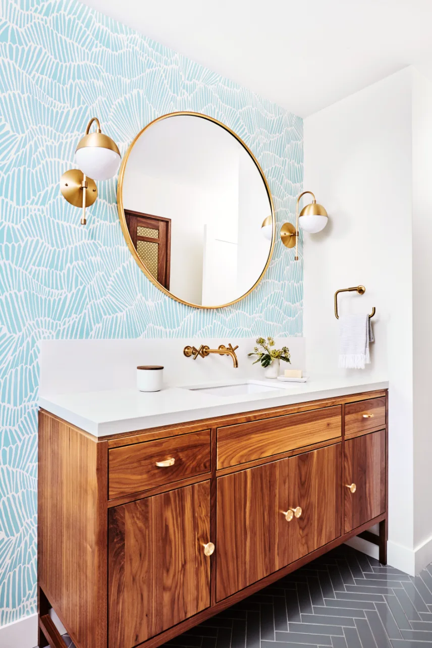 A wooden bathroom vanity with a white countertop is set against teal patterned wallpaper with a round gold mirror and brass sconces.