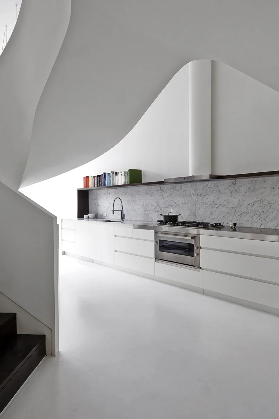 A minimalist white kitchen features handleless cabinets beneath a sweeping, curved white plaster ceiling and a veined marble backsplash.