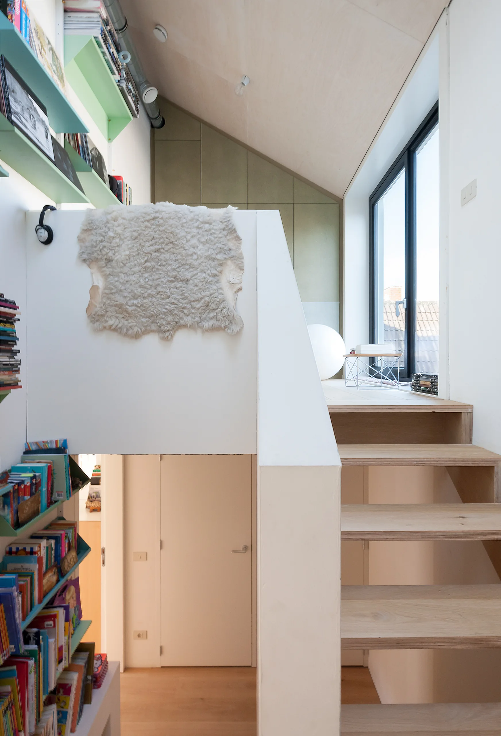 A wooden staircase leads to a loft landing with a sloped plywood ceiling, a large window, and green built-in bookshelves.