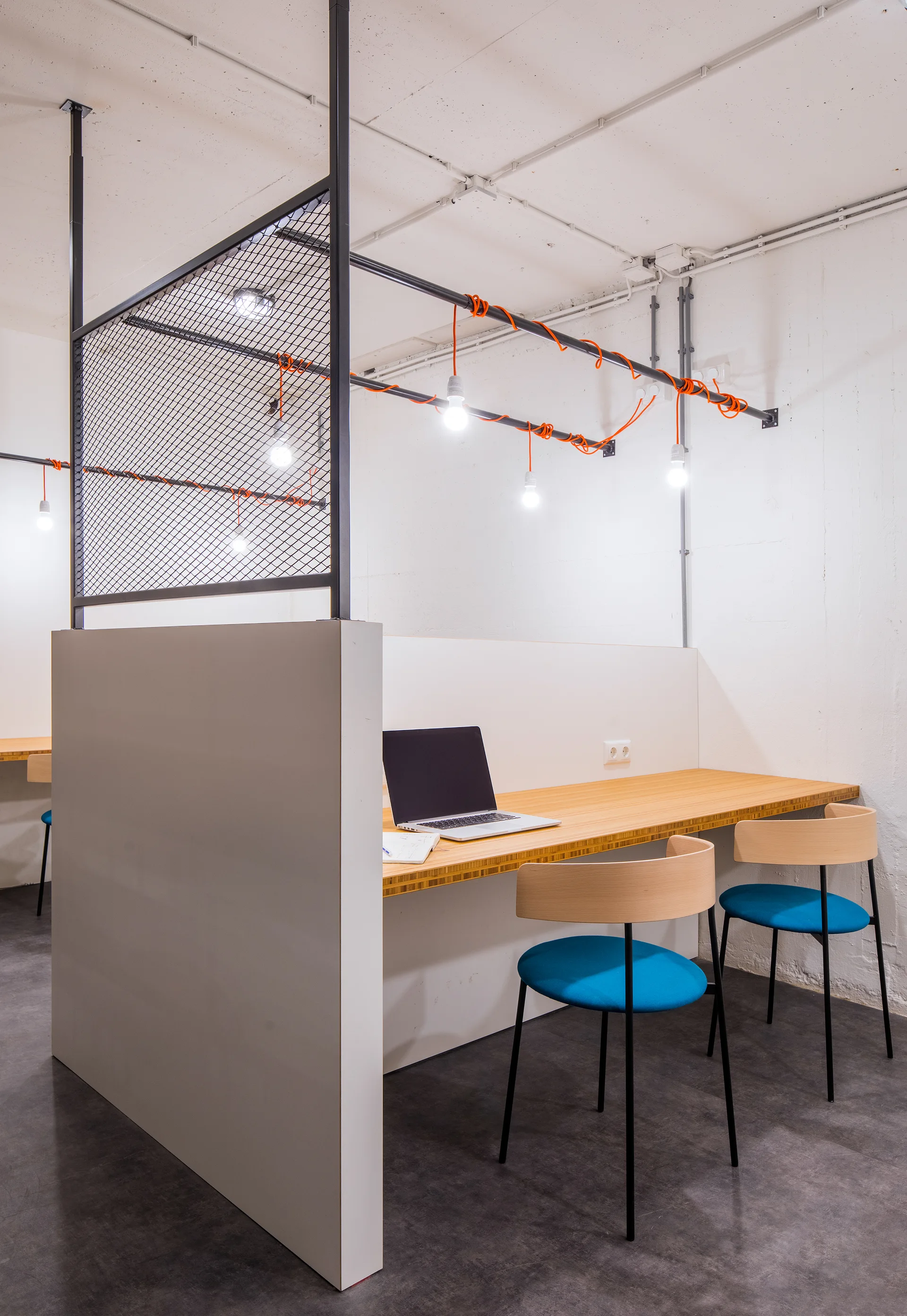 A modern workspace features a light wood desk, blue chairs, a grey partition with black mesh, and exposed bulb pendant lights.