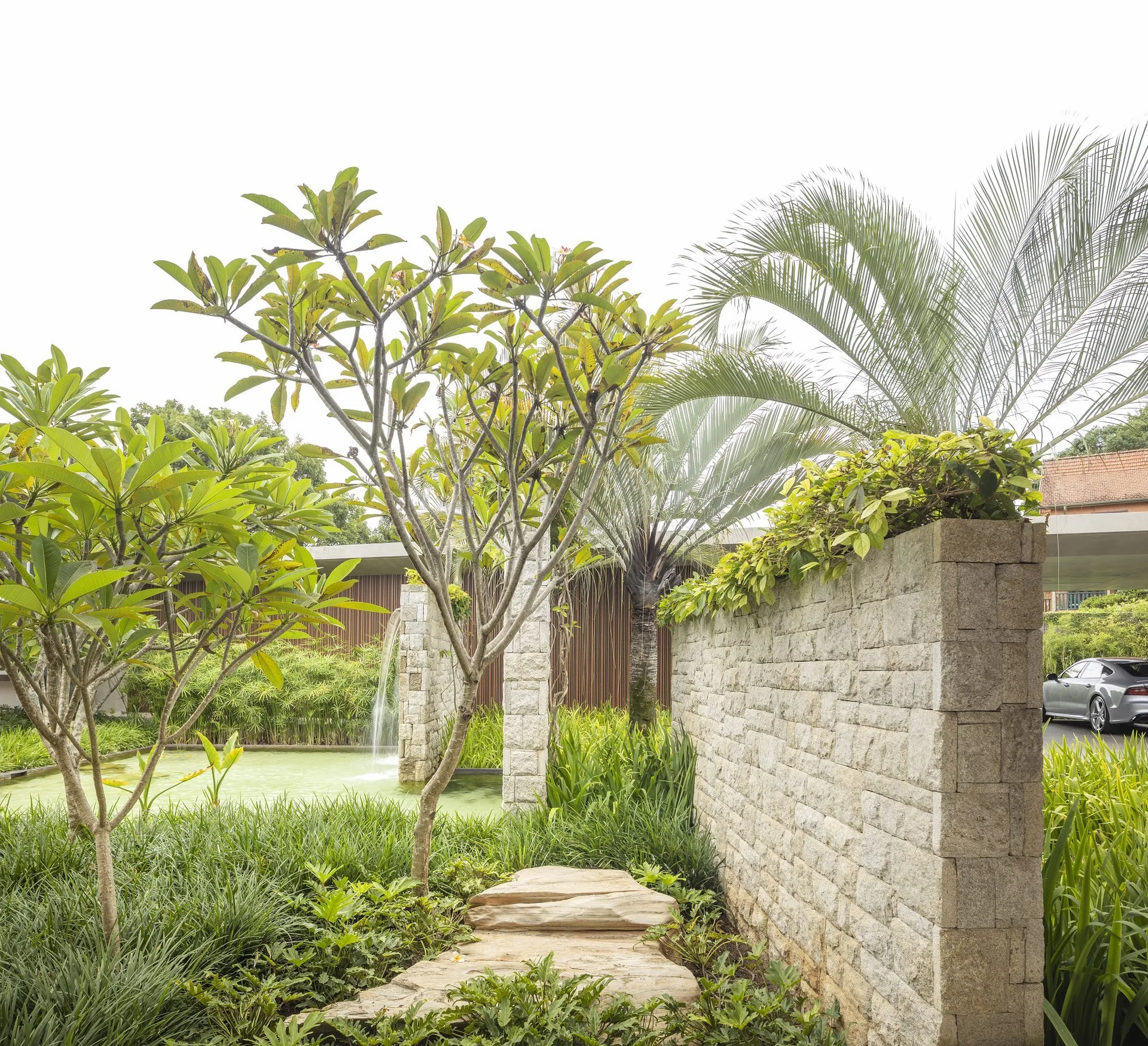 A stone pathway leads past a textured stacked stone wall toward a water fountain surrounded by lush green tropical plants.