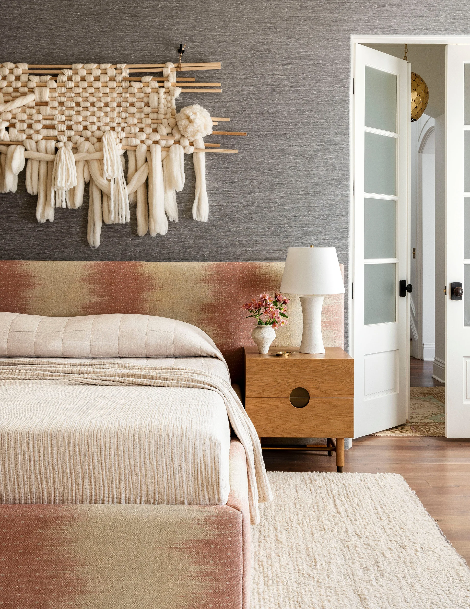 A bedroom features a pink patterned bed against grey textured wallpaper, topped with a large woven macrame hanging next to a wooden nightstand.